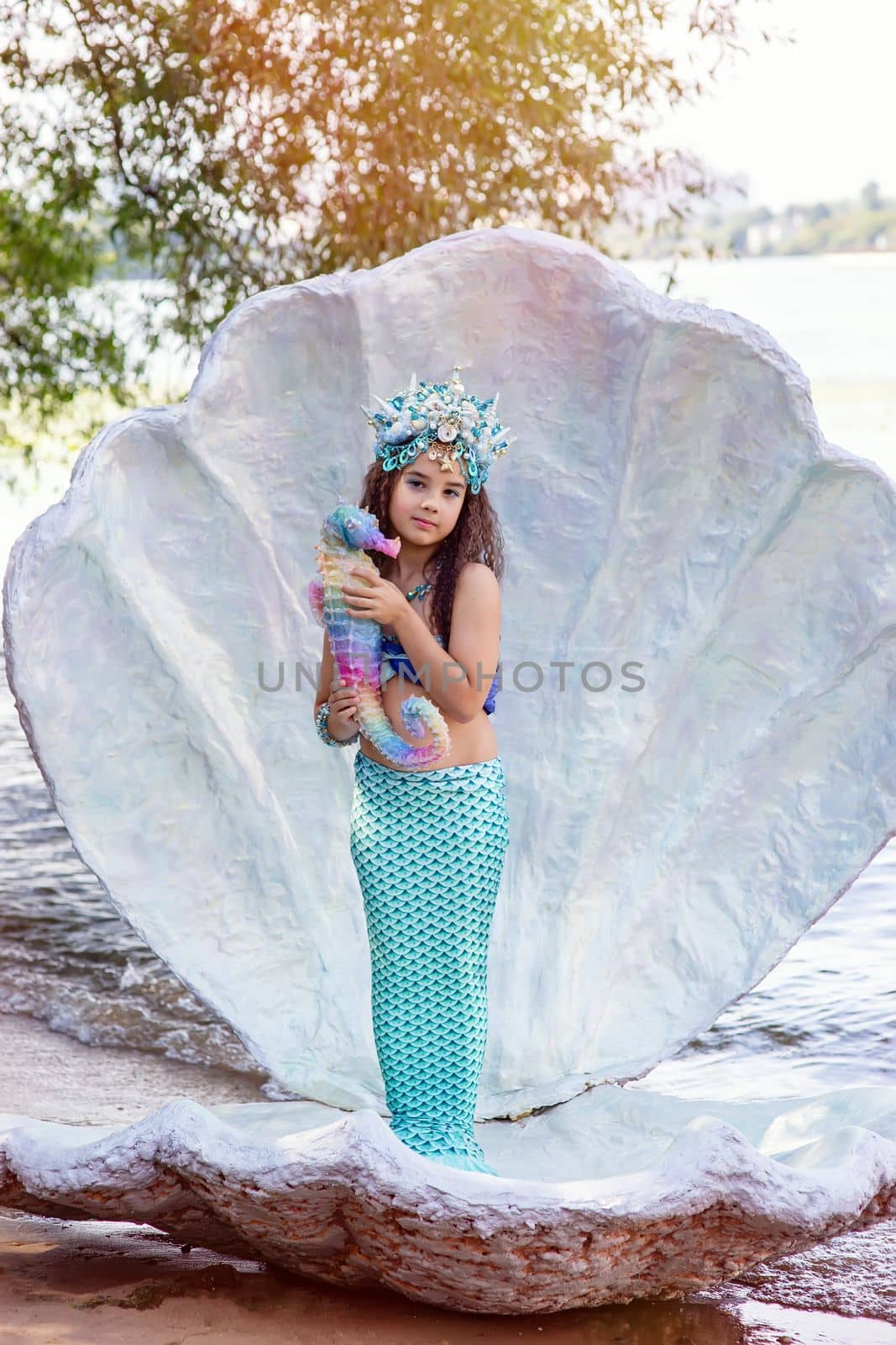 A smiling little girl in a turquoise mermaid costume and a crown is standing outdoors, in a large seashell, holding a colorful seahorse toy. Vertical. Close up. Copy space