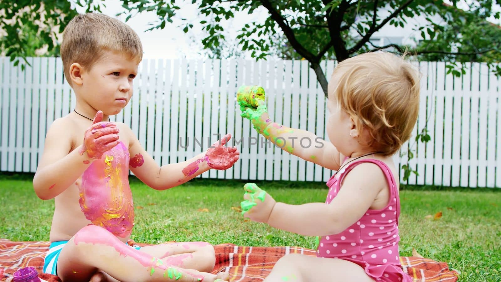 small children, a four-year-old boy and a one-year-old girl, brother and sister, play together, paint with finger paints, in the garden, sitting on a blanket, on grass, lawn, in summer. High quality photo