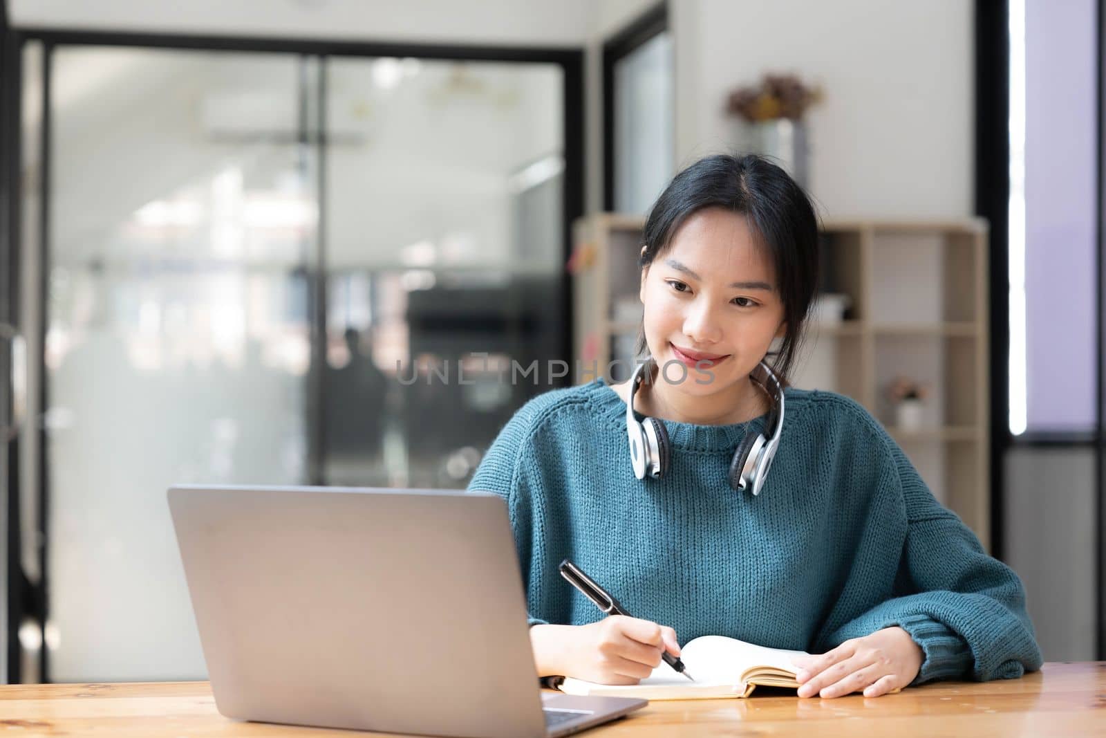 Young adult happy smiling Asian student wearing headphones talking on online chat meeting using laptop in university campus or at virtual office. College female student learning remotely