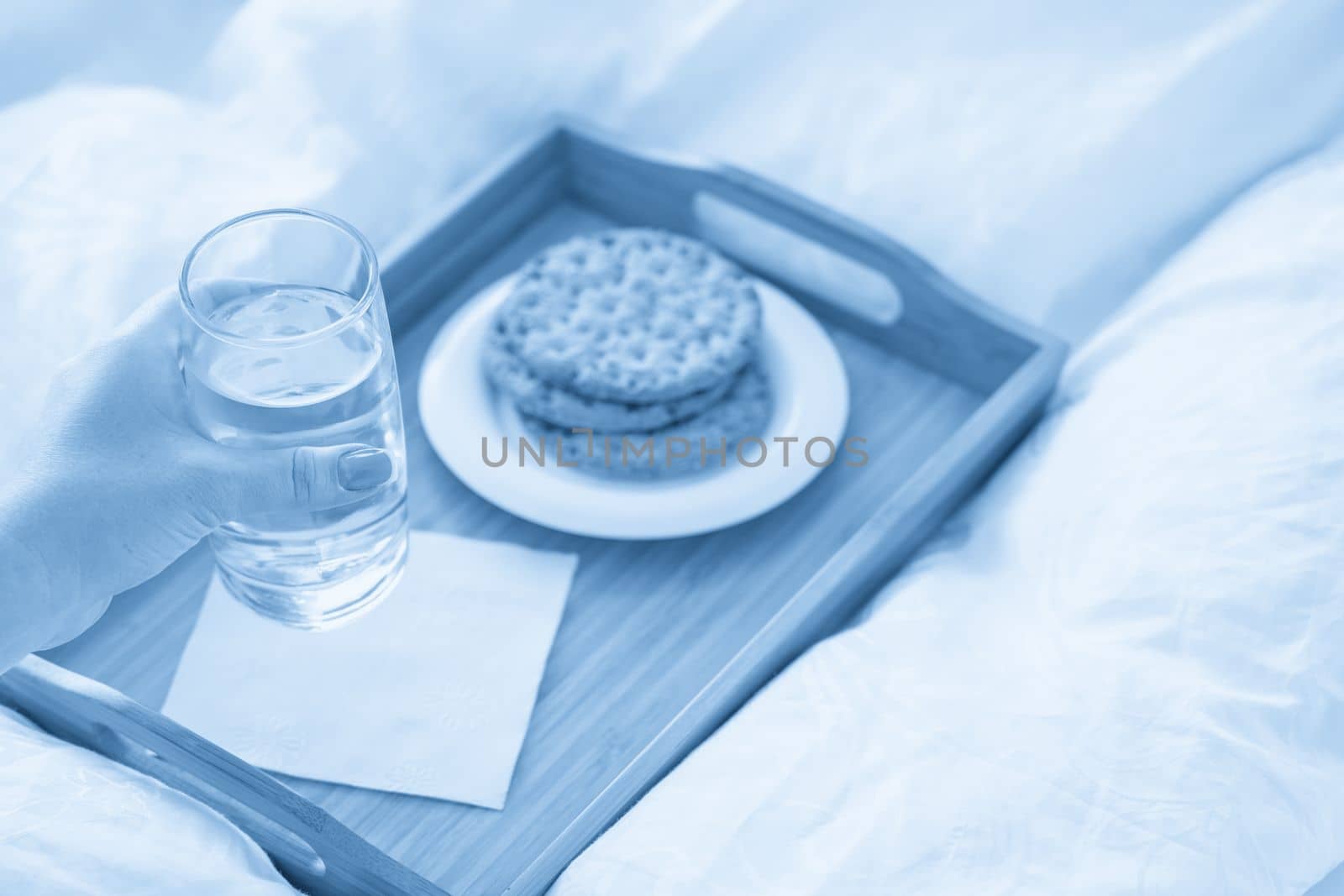 Female hand holding glass of water over tray with crackers on bed by Mariakray