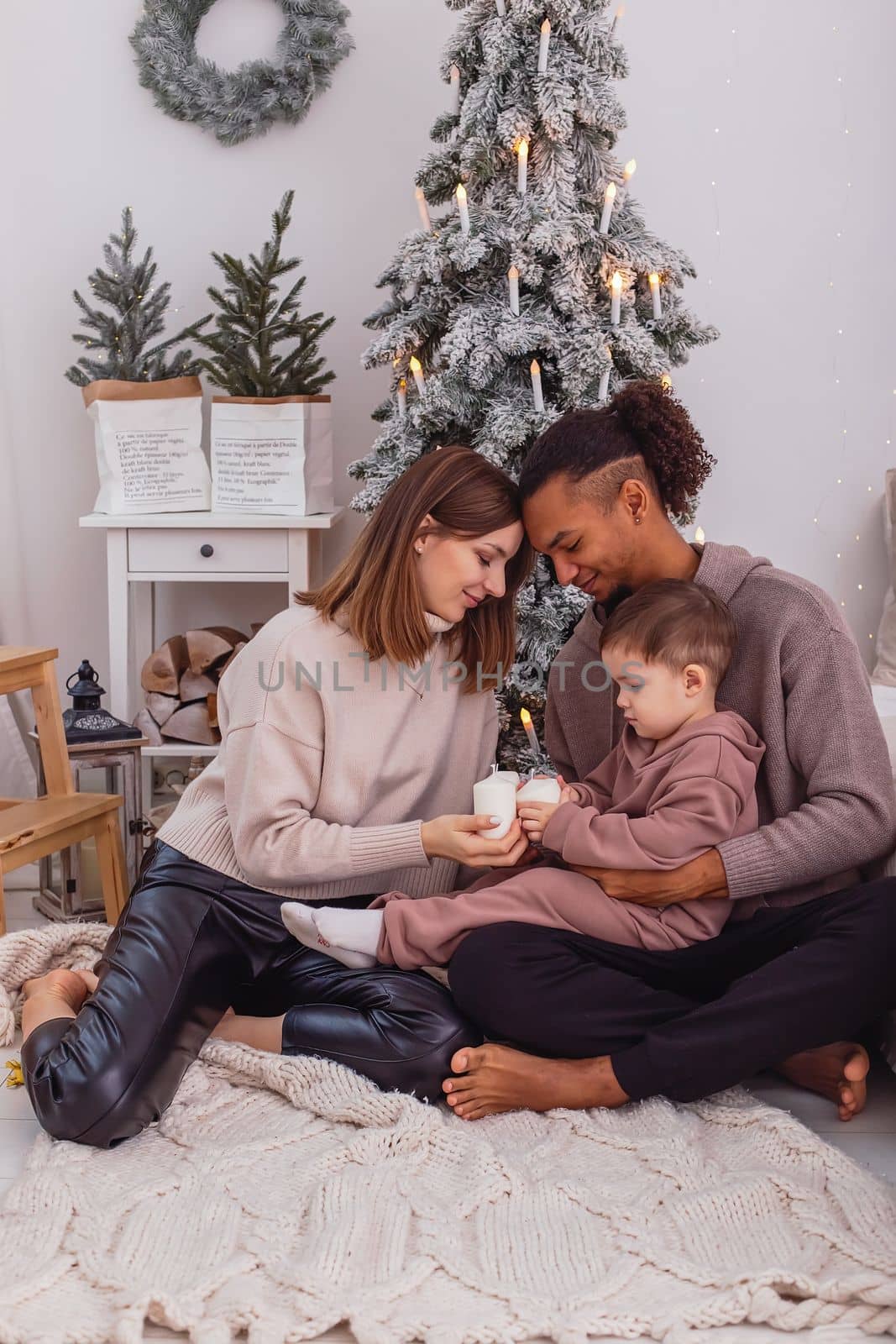 A young multi-racial family with a child is sitting on a knitted blanket near a Christmas tree by Zakharova