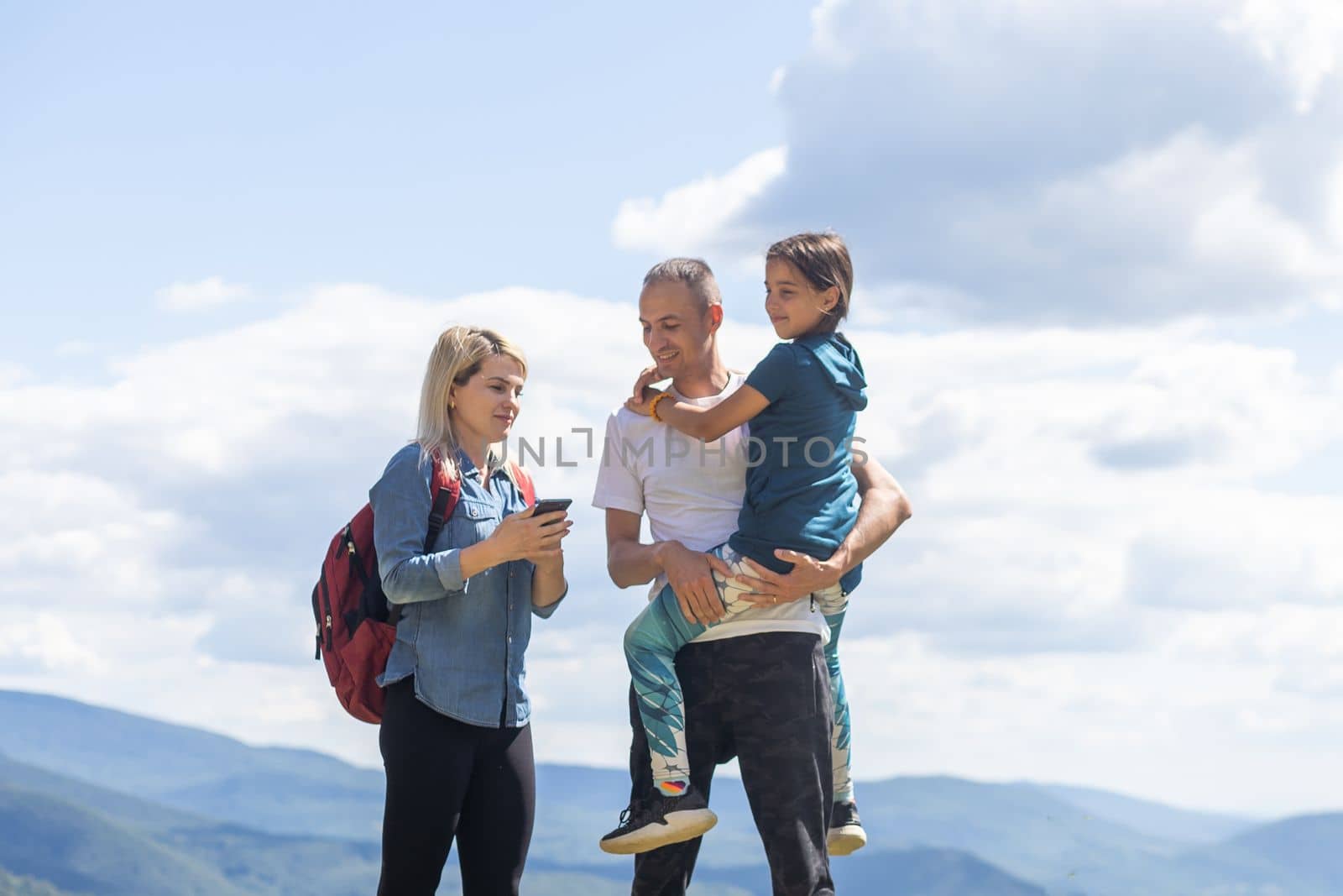 Mother, father hugging son, and daughter in summer mountains. Happy family smiling and standing on the green grass in the field at sunset. Kids with parents. Family weekend.