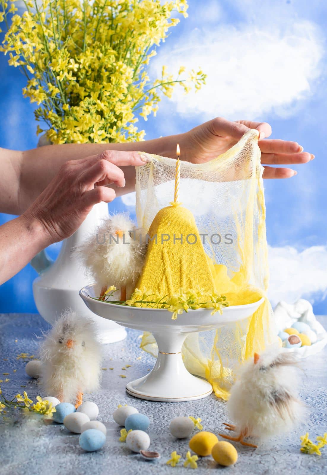 a woman lights a candle on a traditional yellow Slavic Easter curd cake on a blue background against blue sky with white clouds, High quality photo