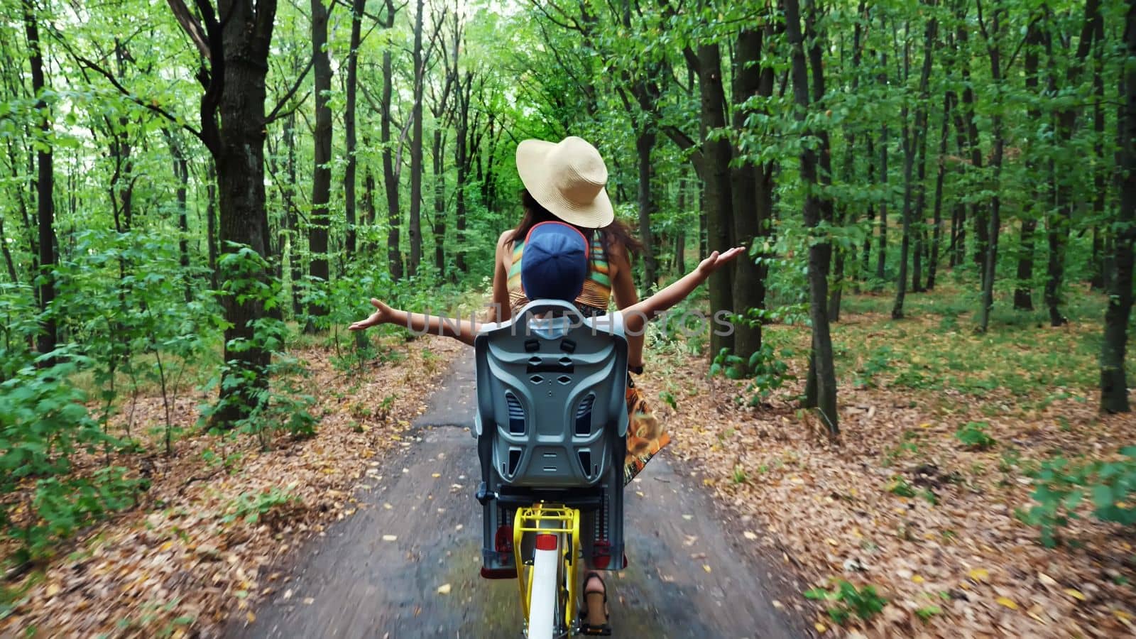 a woman in a hat and dress, with a basket of flowers, together with small child, ride bicycle, in the forest, in the summer. the child is sitting in a special chair. High quality photo