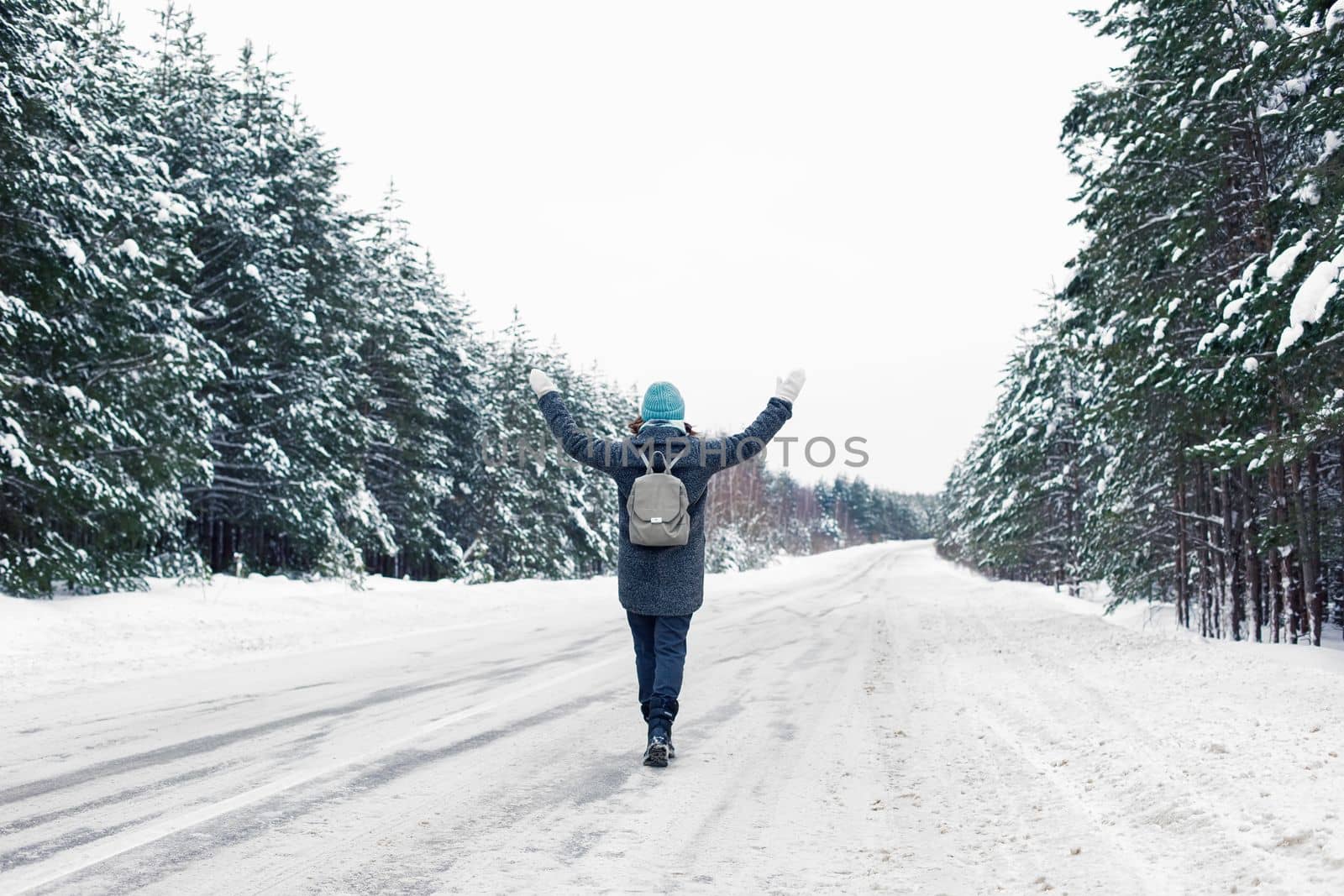 a girl in a gray coat and a blue knitted hat, with a gray backpack on her back, walks along the side of a winter road, raised her hands up. Nearby there is a beautiful winter pines forest. Back view