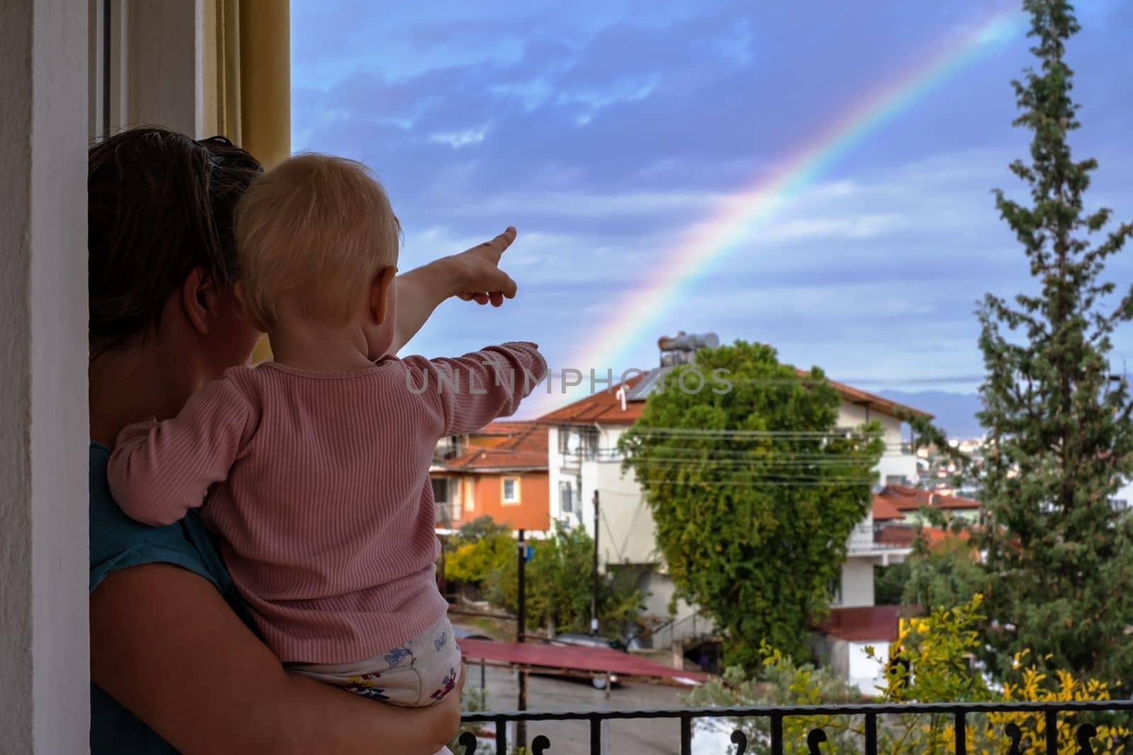Mother looks at the sky with a baby in her arms, admires the rainbow after the rain, summer is outdoors. 