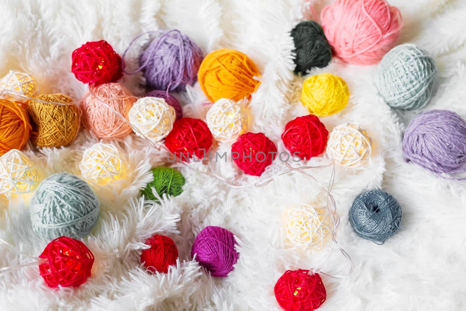 Bright multicolored knitting yarn and a luminous garland in the form of balls lie on a white fluffy blanket. Cozy still life made of yarn. Close up.