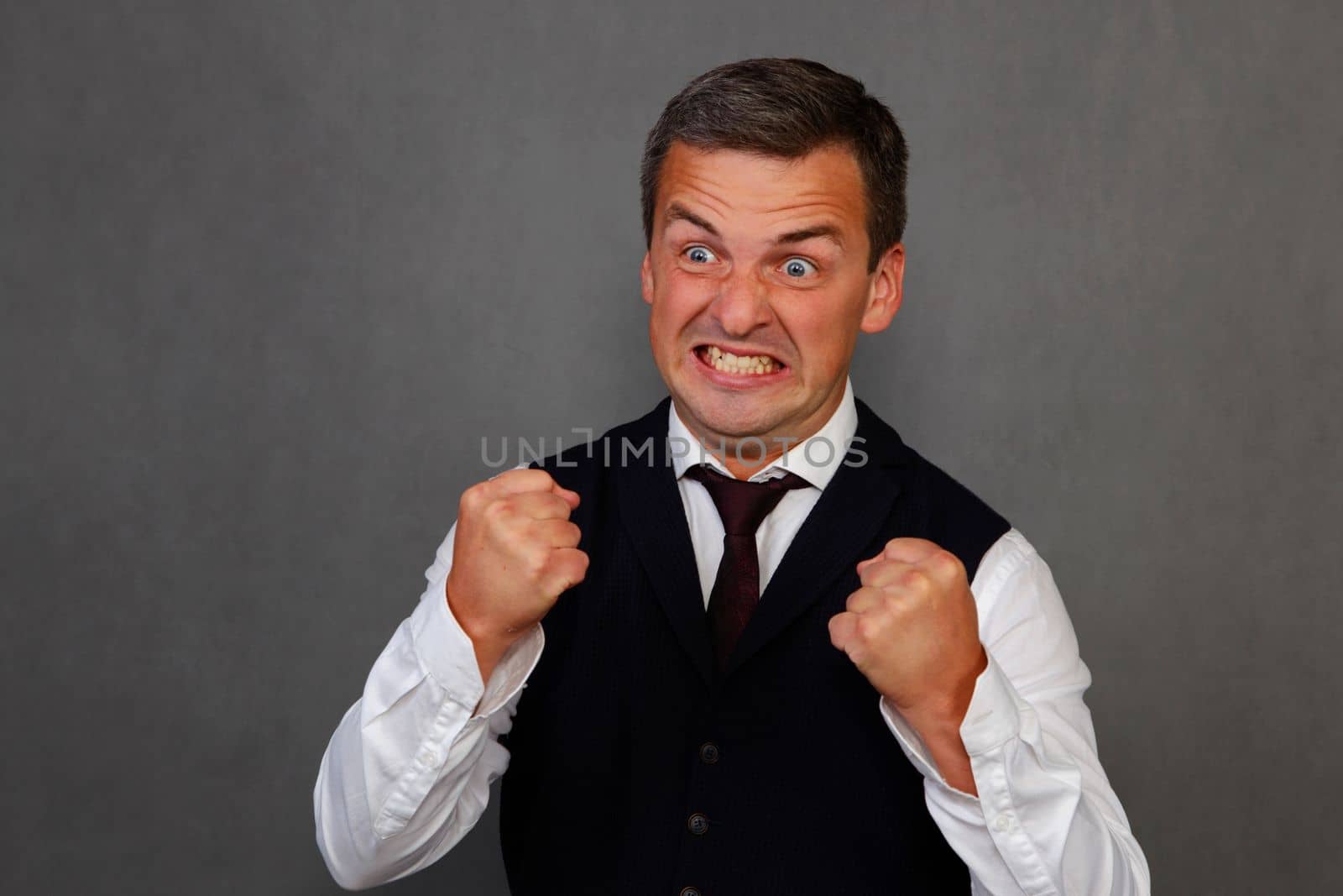 A handsome man in a white shirt, tie and dark waistcoat on a gray background got very angry. The person is in a state of intense rage.