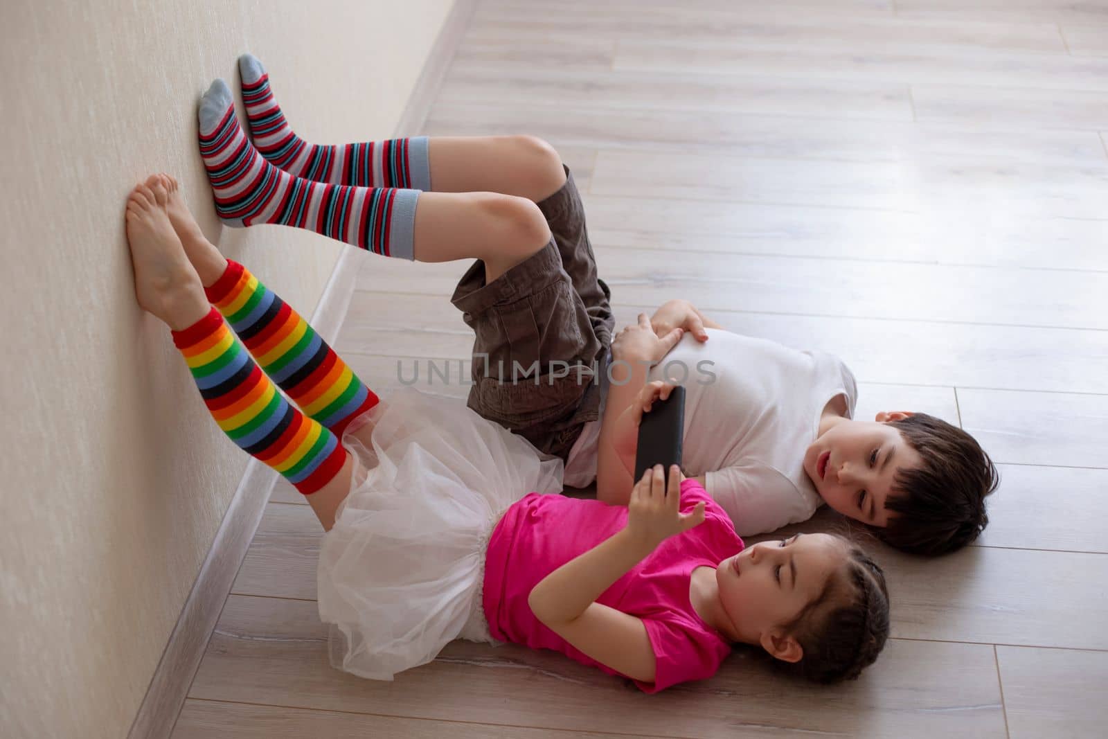 Two children, brother and sister, are lying on the floor with their legs in bright striped leggings on the wall and playing on a smartphone. Stay home while quarantined