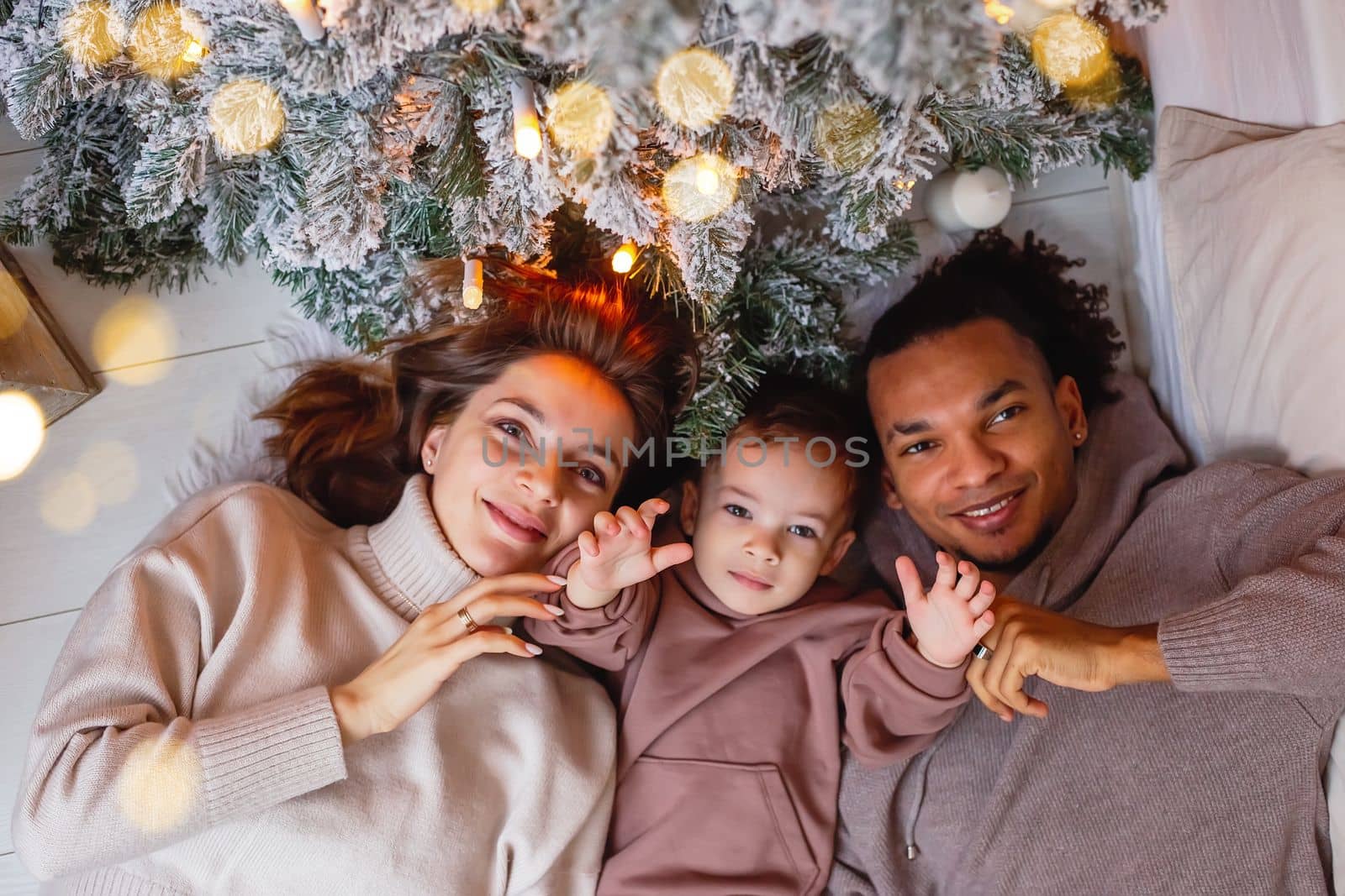 A happy young multiracial family with a little boy are lying on a knitted blanket, under a Christmas tree. Top view. Close up