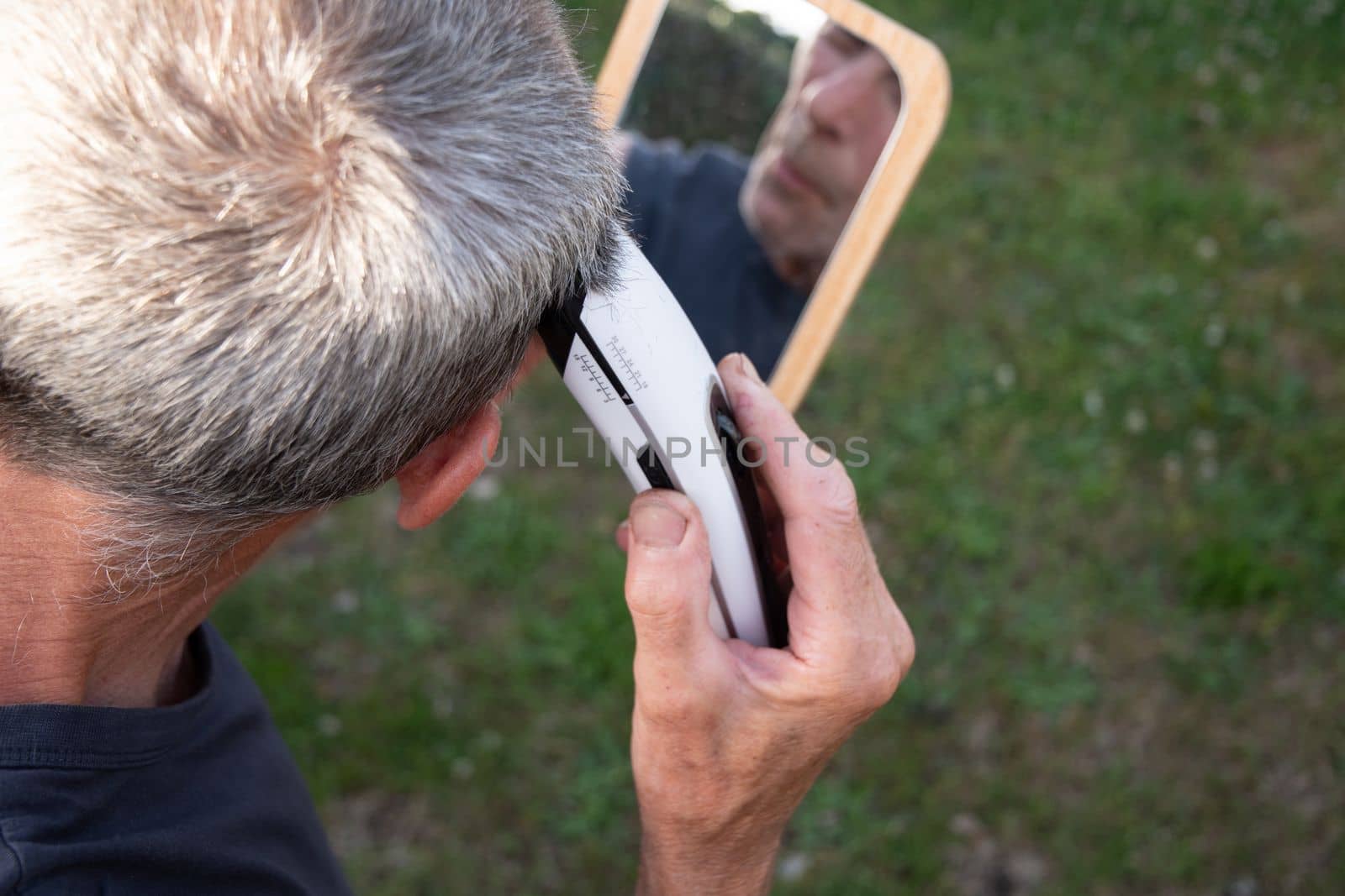 a gray-haired middle-aged man shaves his hair with a clipper in a garden on a green lawn and looks into a table mirror. High quality photo