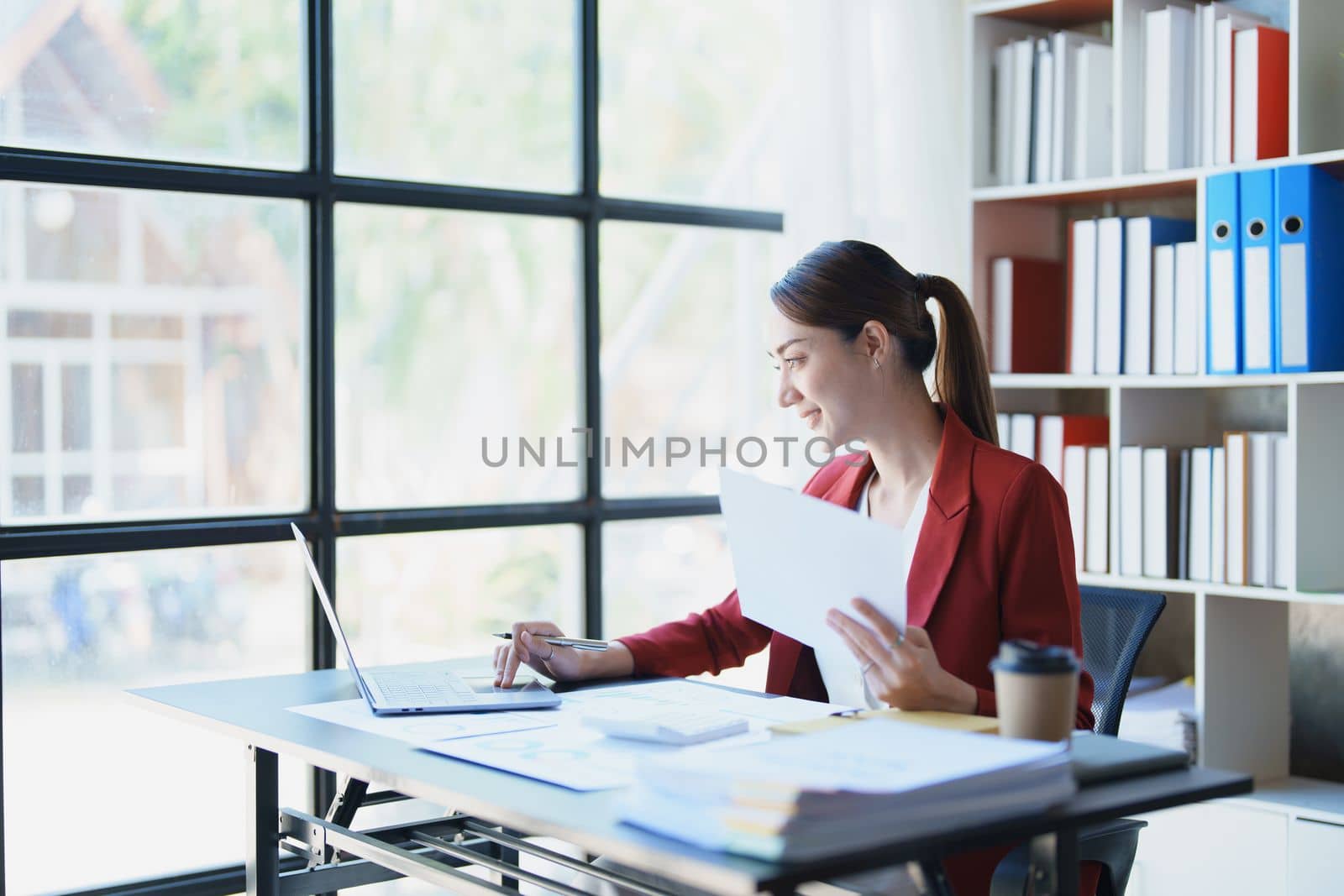 Portrait of a thoughtful Asian businesswoman looking at financial statements and making marketing plans using a computer on her desk by Manastrong