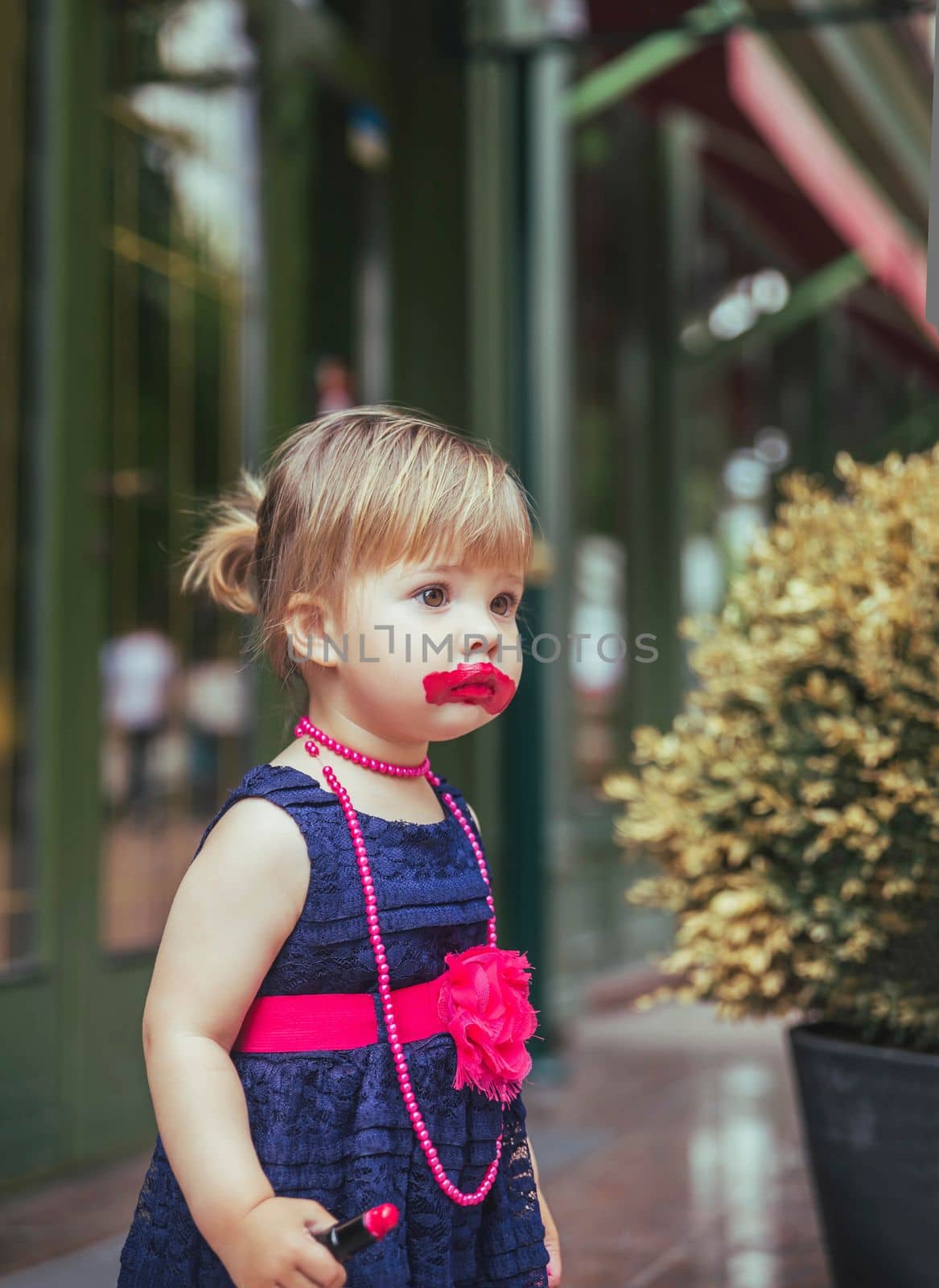 Adorable baby in a dress paints lips with lipstick.