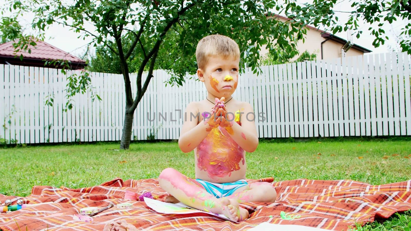 a little child, a four-year-old boy playing, painting with finger paints, decorating himself, in the garden, sitting on a blanket, on grass, lawn, in the summer. he's having fun. High quality photo