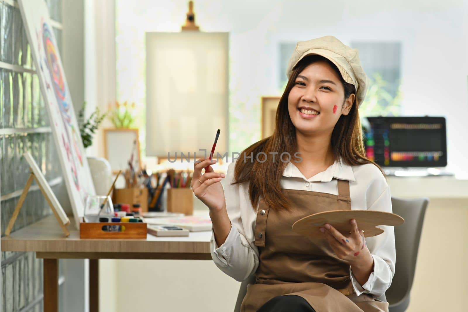 Pleasant female artist in apron with palette and brush painting on canvas at art studio. Education, hobby, art concept.