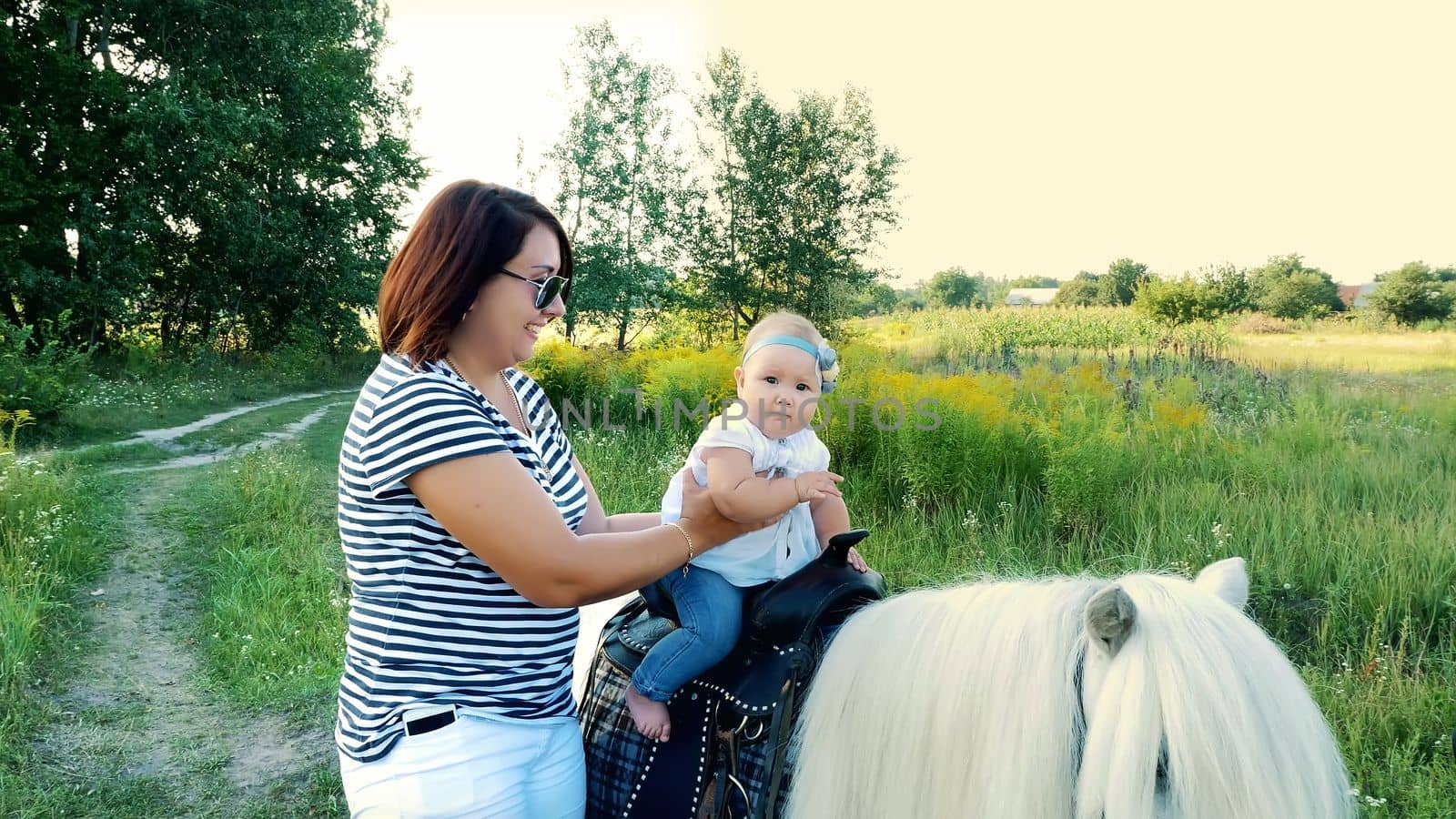 A woman with a baby walks around the field, a baby sits on a pony, mom holds the baby. Cheerful, happy family vacation. Outdoors, in the summer, near the forest. High quality photo