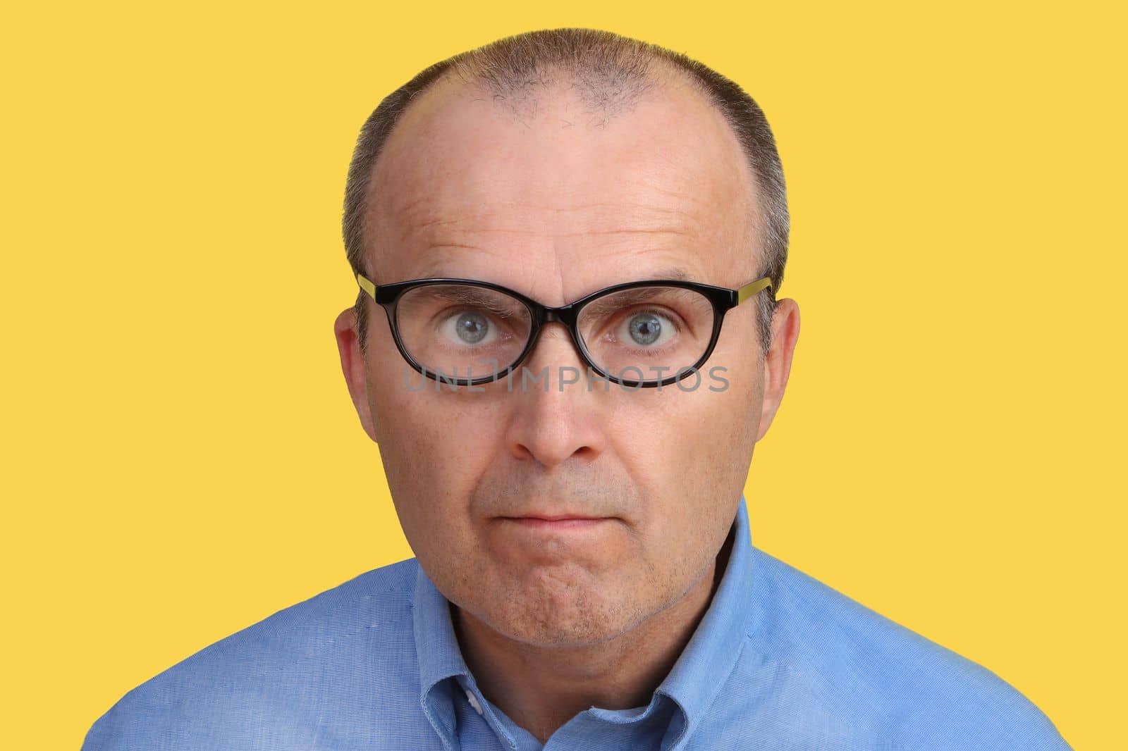 A man with a funny look in glasses and a blue shirt on a yellow background. The man with the glasses looks at the camera.