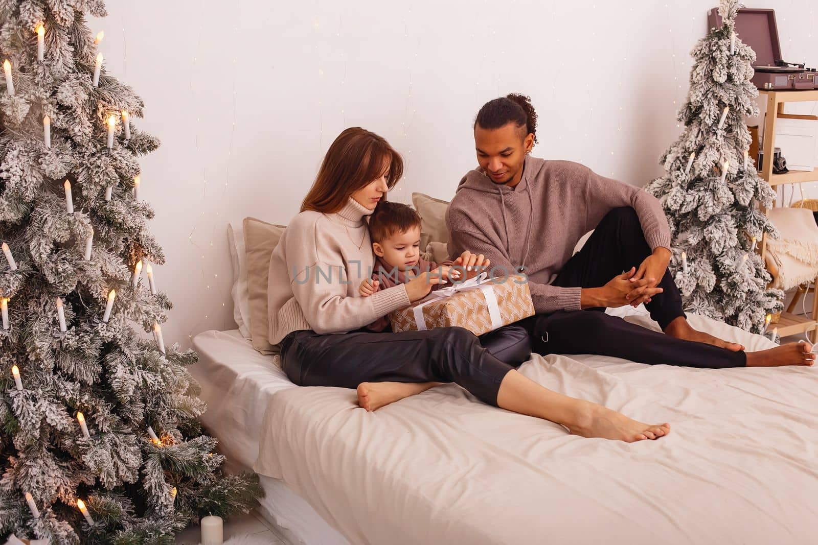 A happy family is sitting on a bed, in a Christmas interior, untying a ribbon on a brown gift box by Zakharova