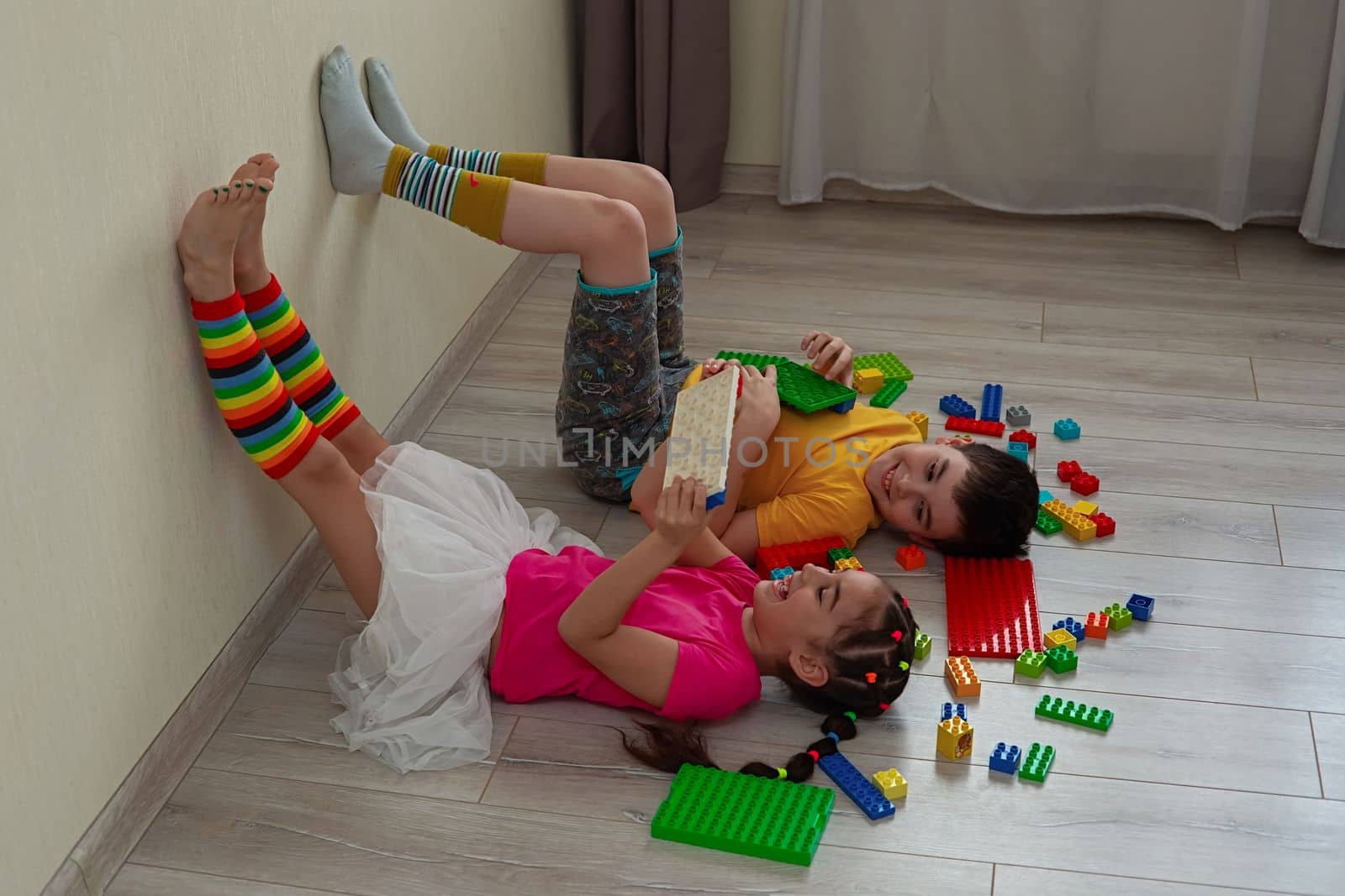 A happy boy and a little girl, brother and sister, brunettes, in bright multicolored clothes, lie on the floor, on their backs, in the room, play merrily with a multicolored plastic construction set