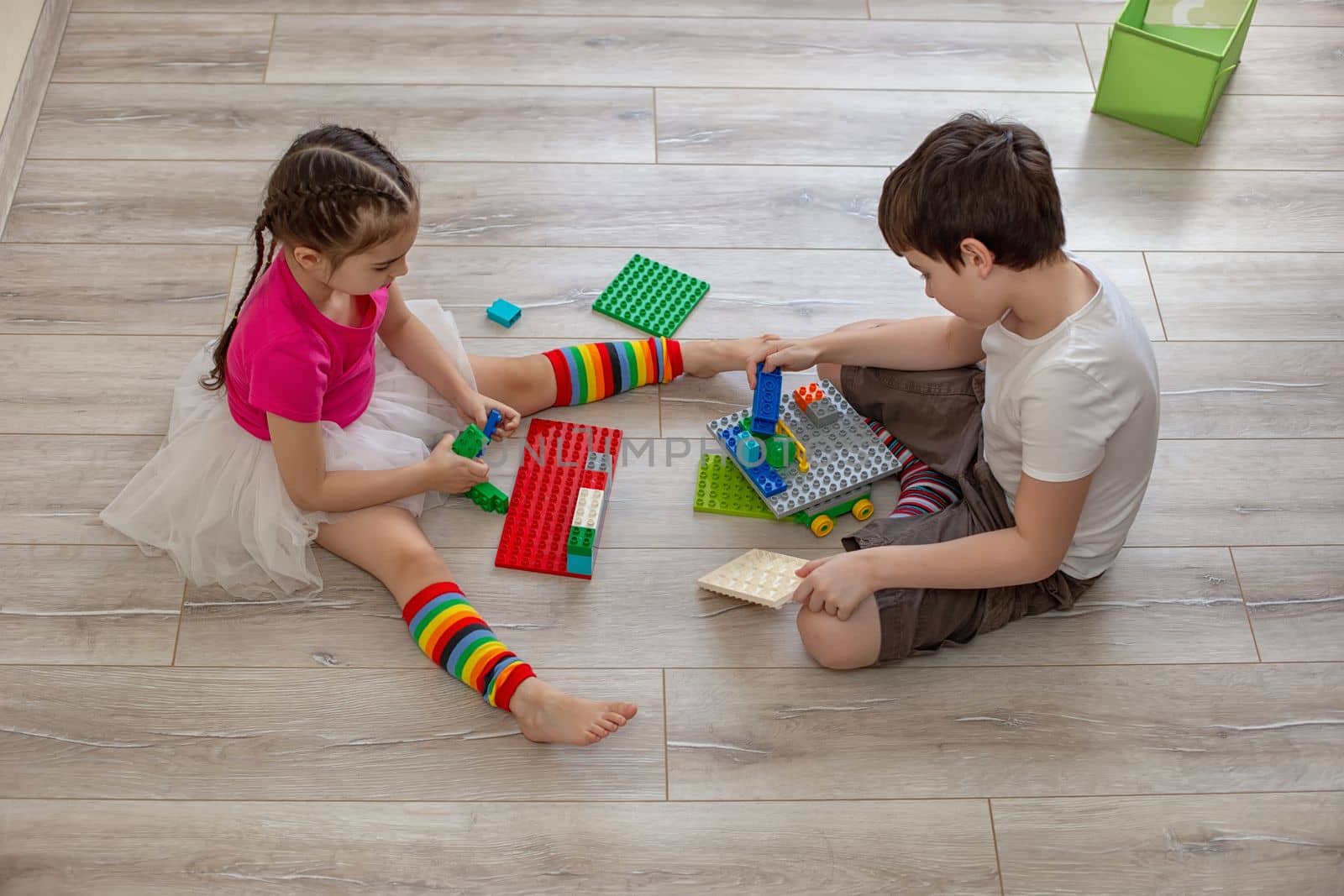 Two children, a boy and a girl, sit on the floor in room and play in plastic constructions. Top view
