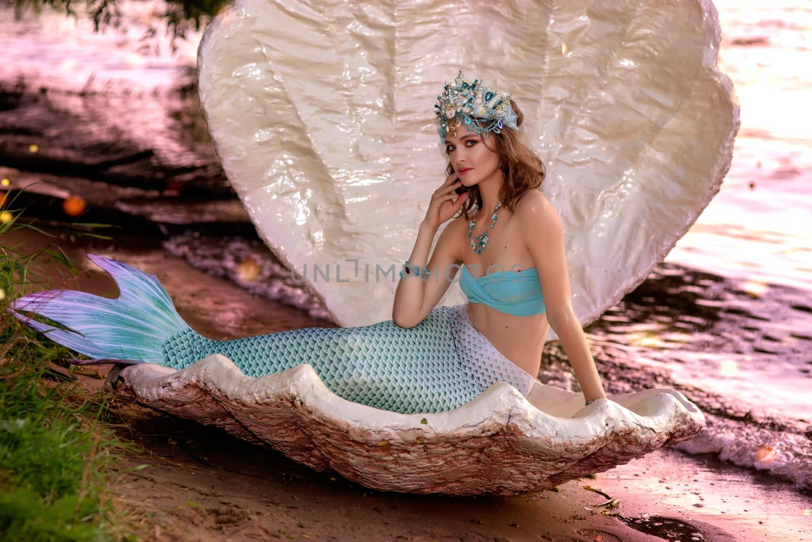 A mermaid girl in a beautiful diadem sits by the water in a large white shell