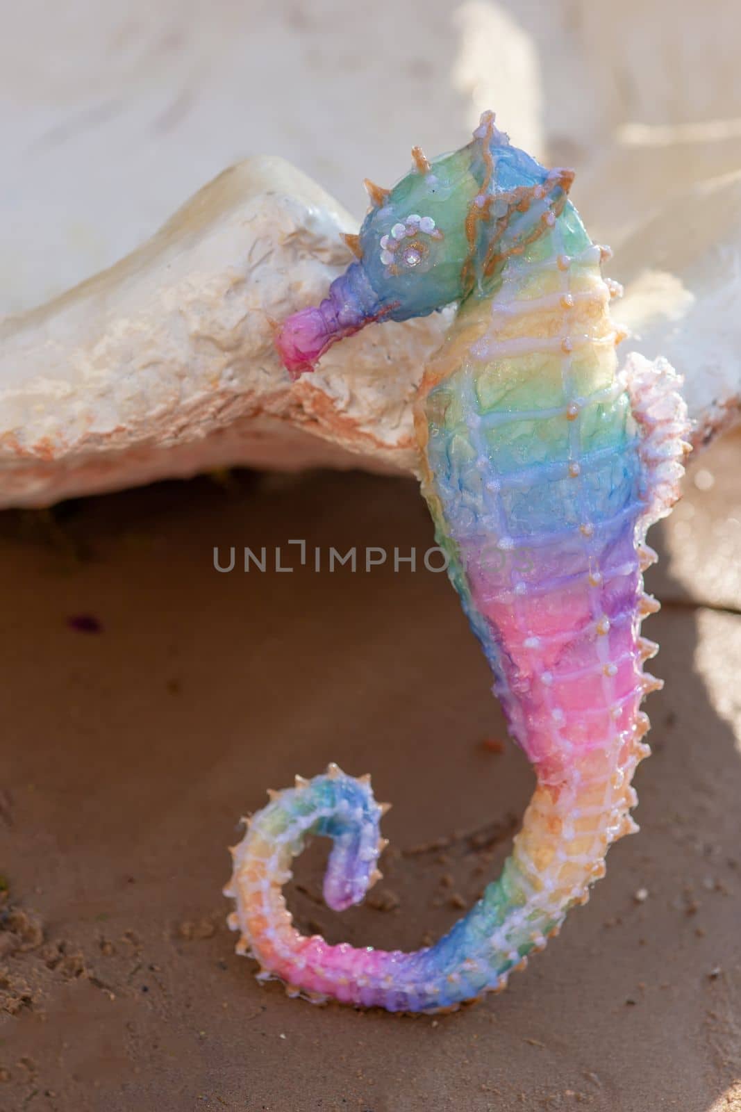 a decorative rainbow seahorse stands on the beach on a sunny day, near a large seashell. close up