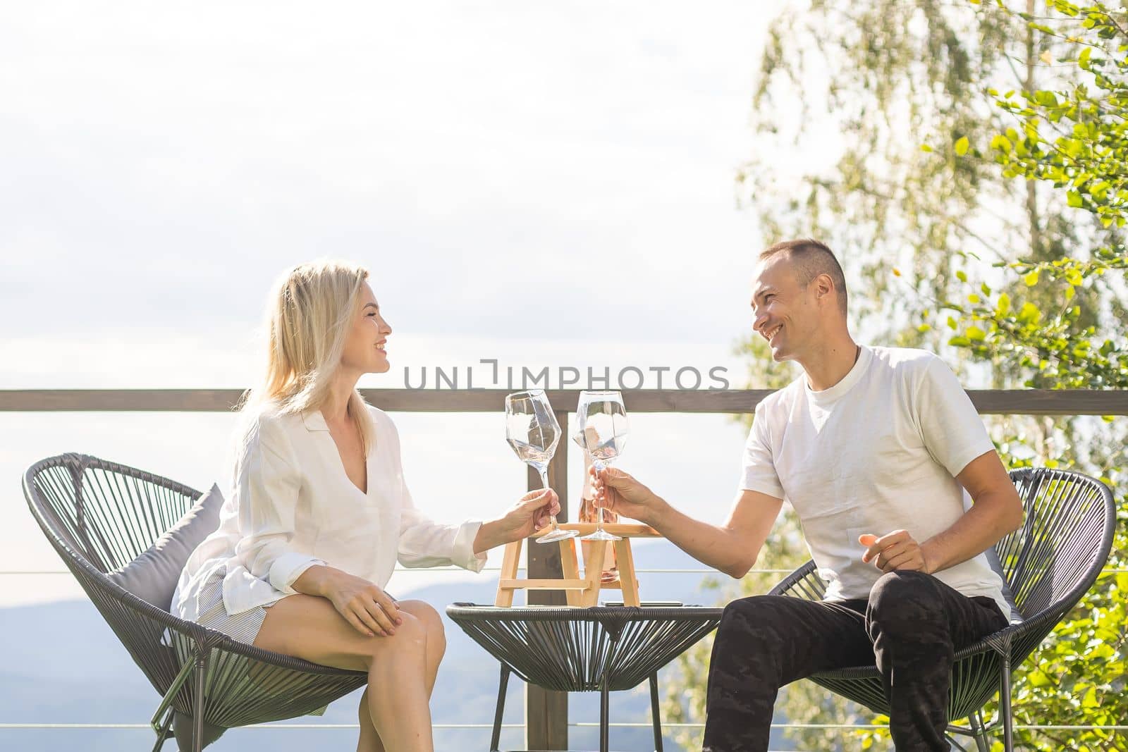 Happy couple sit on wicker chairs at outdoor terrace in the mountains and drink wine. Romantic time together