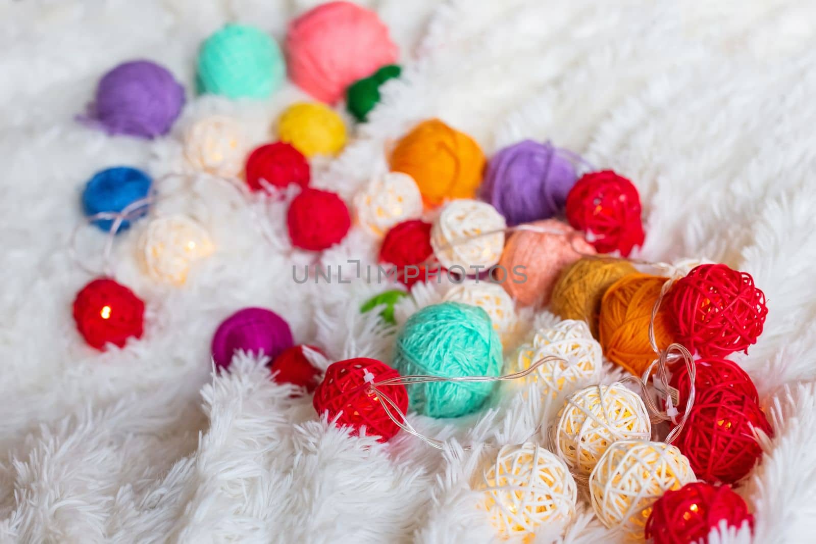 A lot of colorful knitting yarn and a luminous garland in the form of balls lie on a white fluffy blanket. Cozy still life made of yarn. Close up. Copy space