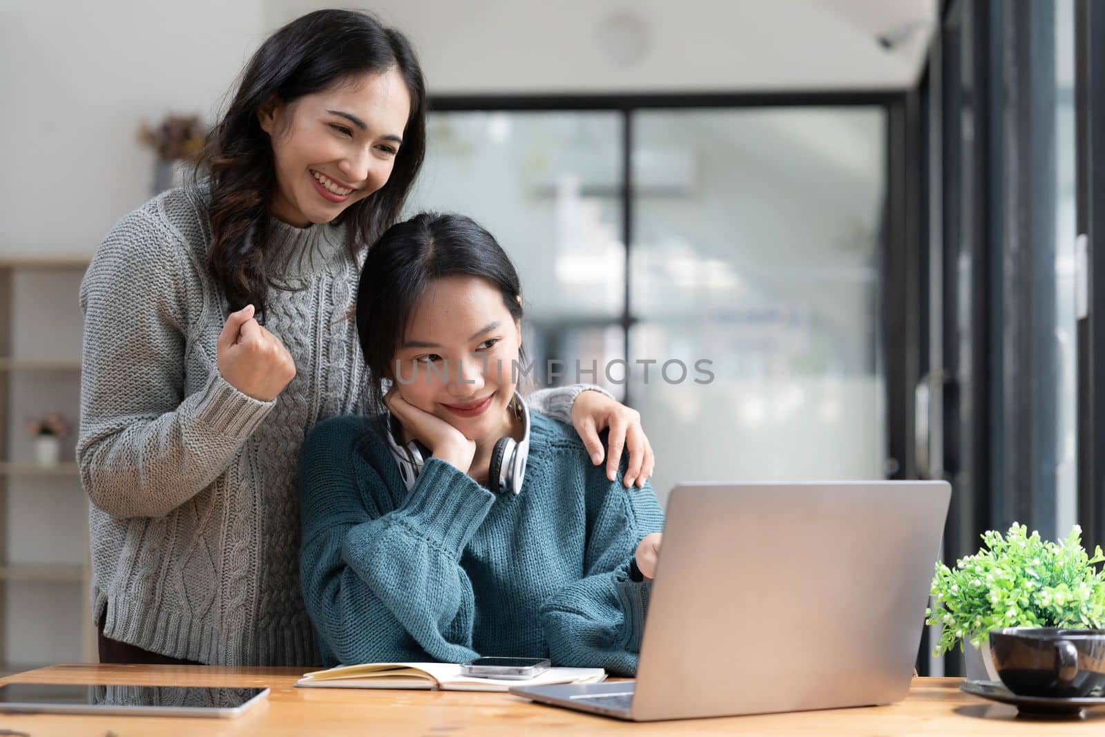 Two young Asian women show joyful expression of success at work smiling happily with a laptop computer in a modern office. by wichayada