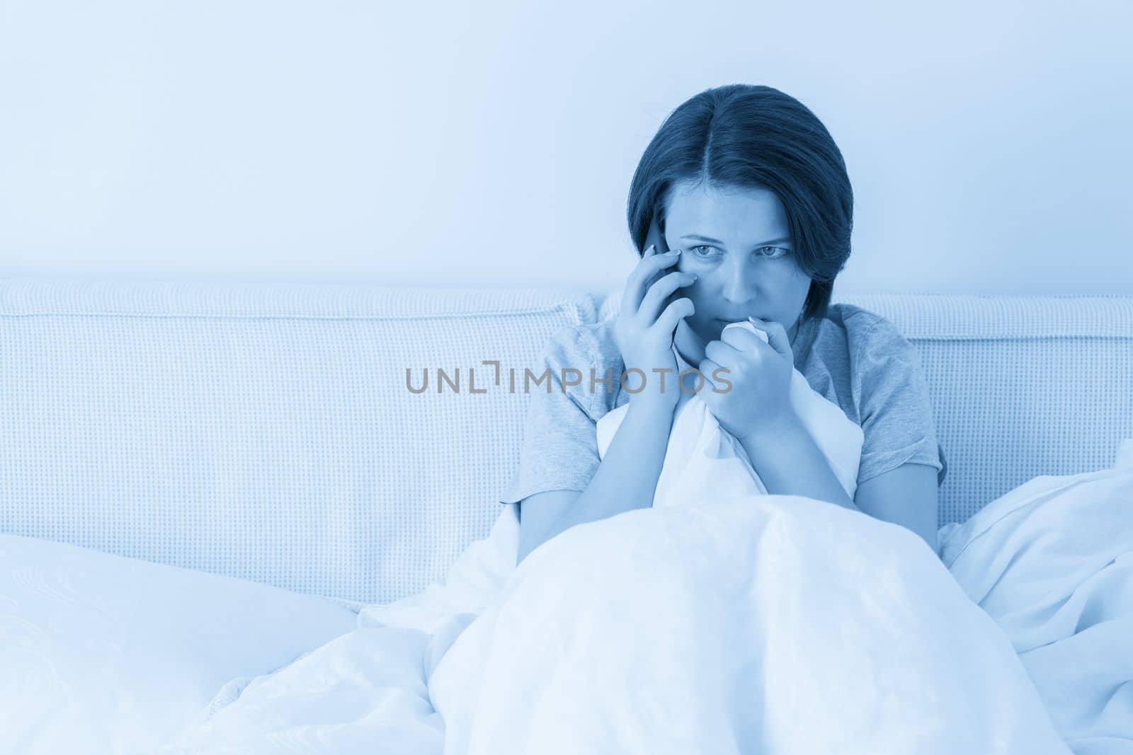 Woman using smartphone as she sits in bed covered with duvet, scared and terrified by Mariakray