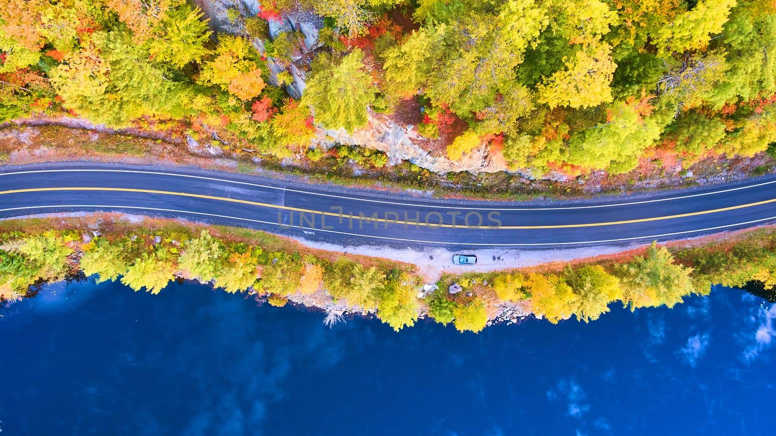Aerial looking down on road between blue water lake and rocky forest in fall by njproductions