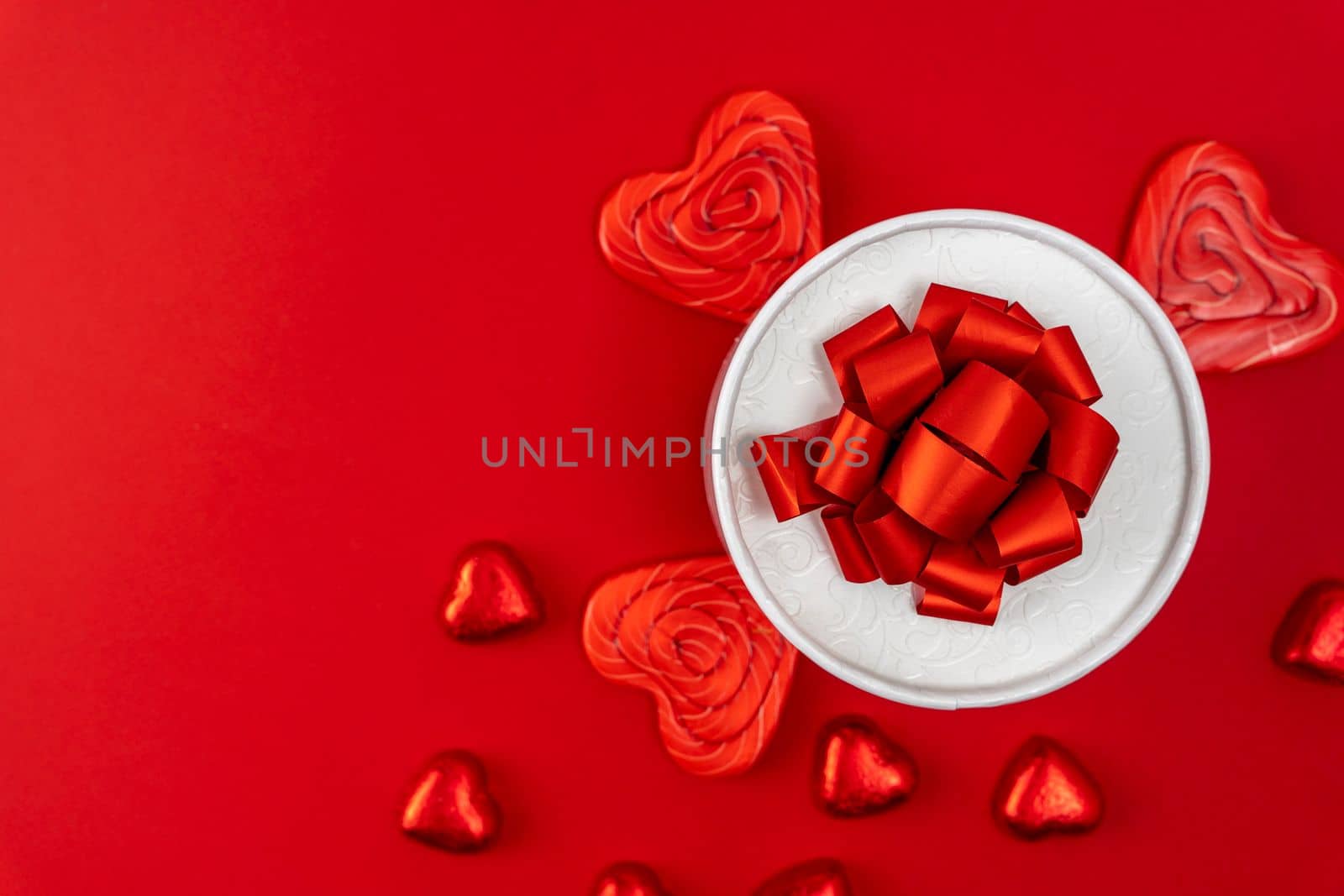 creative minimalist composition. a gift box with a ribbon and a bow, heart-shaped chocolates and heart-shaped caramel lollipops on a red background. A declaration of love. a gift and gratitude. Valentine's Day. postcard. copy space