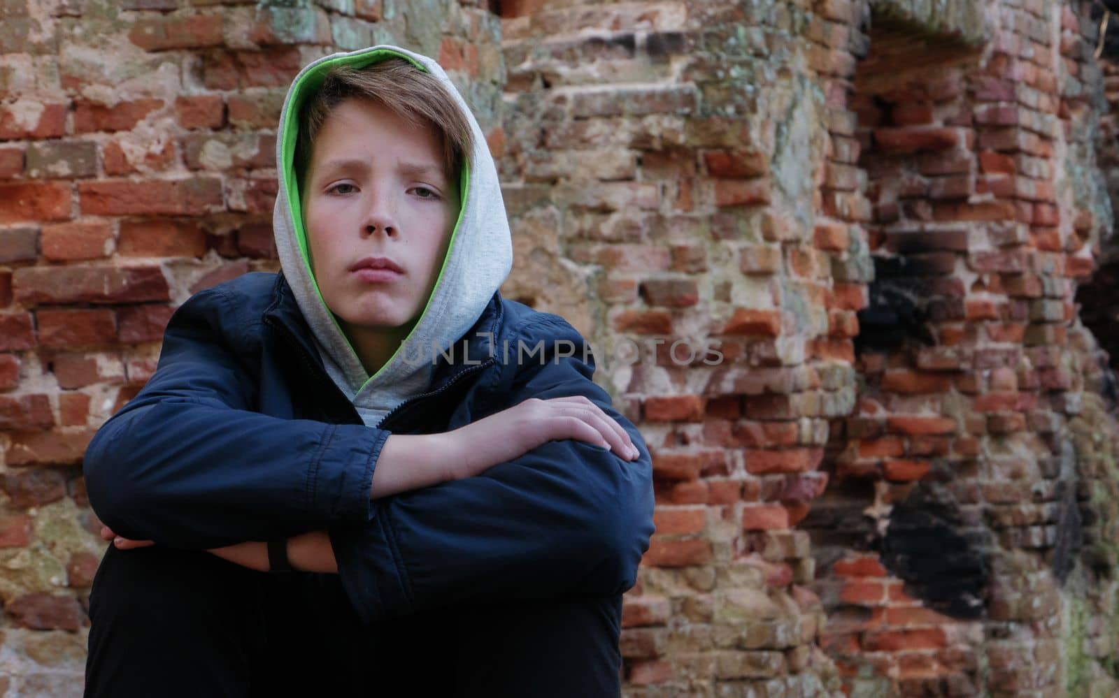 Portrait of a sad cute boy on the background of an old brick wall. A thoughtful boy in a blue hooded jacket looks at the camera against the background of an old brick wall..