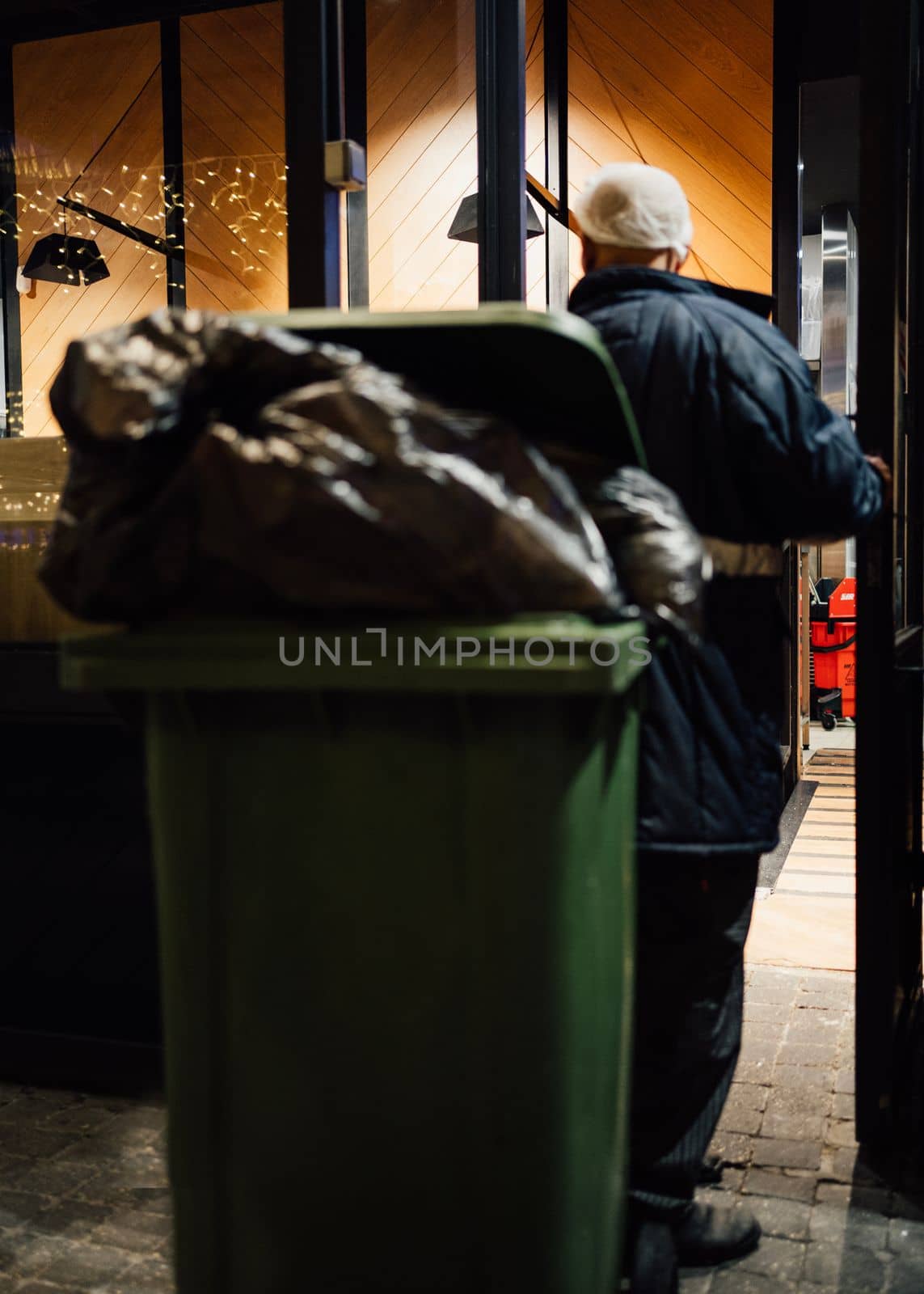 Man worker taking out a loaded green garbage bin waste trash and recycling from bar at christmas. 