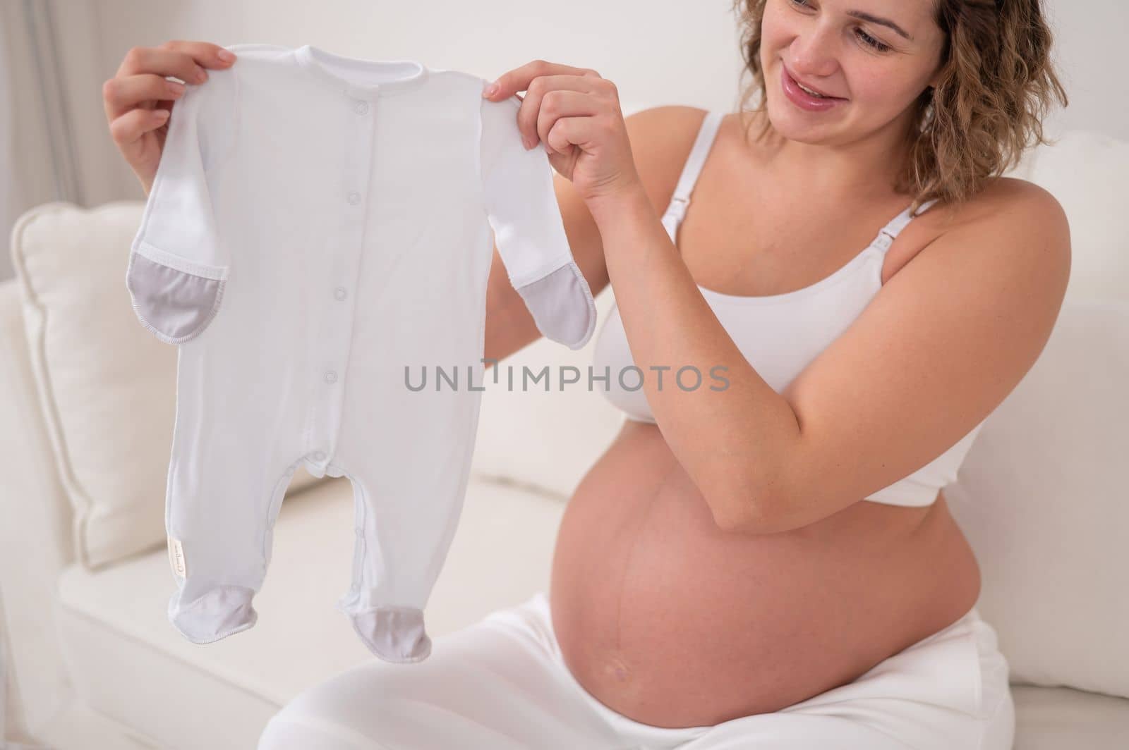 A pregnant woman sits on a white sofa and holds clothes for the expected baby