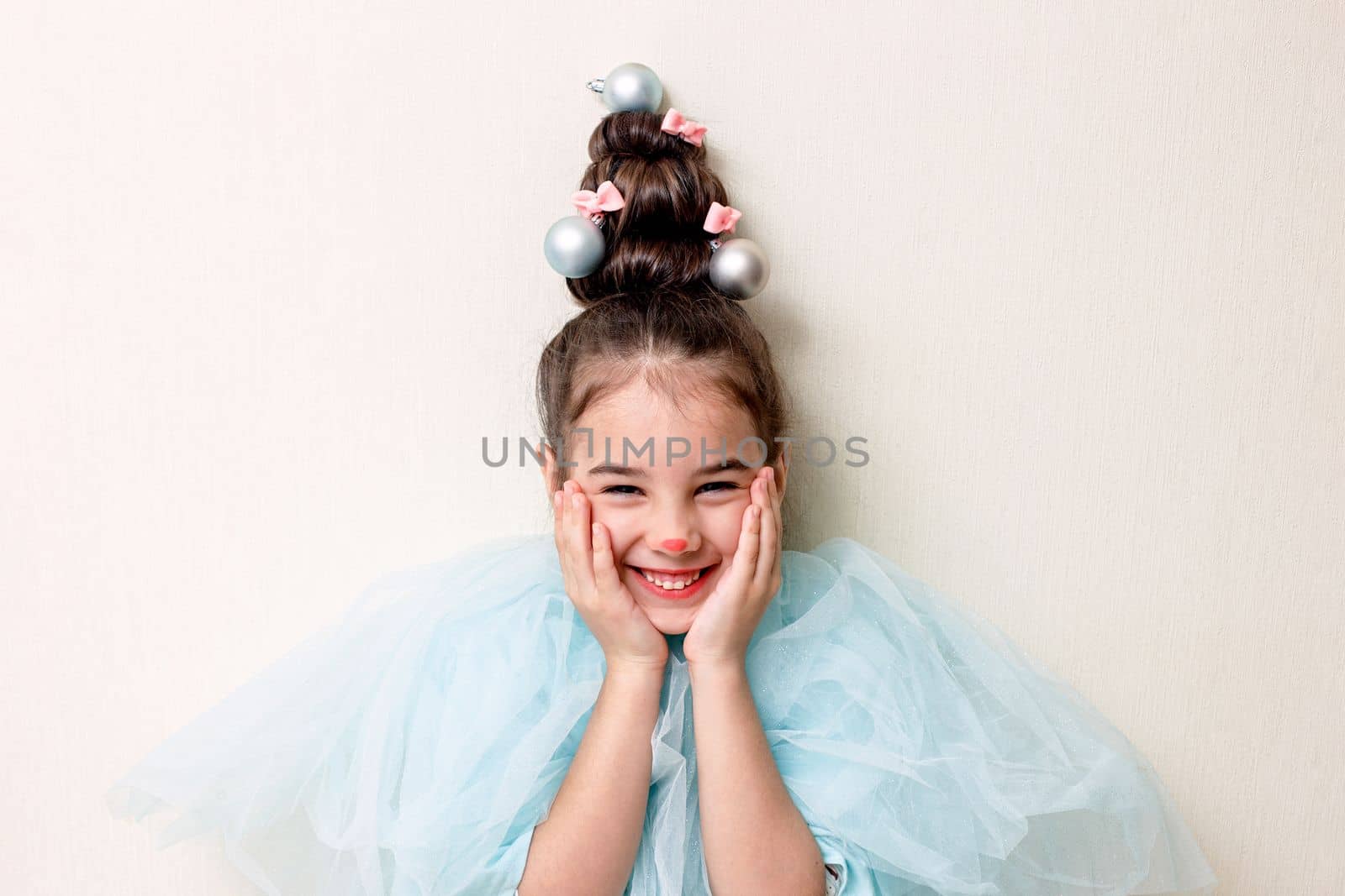 Portrait of a funny beautiful little girl in blue clothes, with a hairstyle in the form of a Christmas tree with a luminous garland, stands against the background of a light wall, pressed her palms to her cheeks