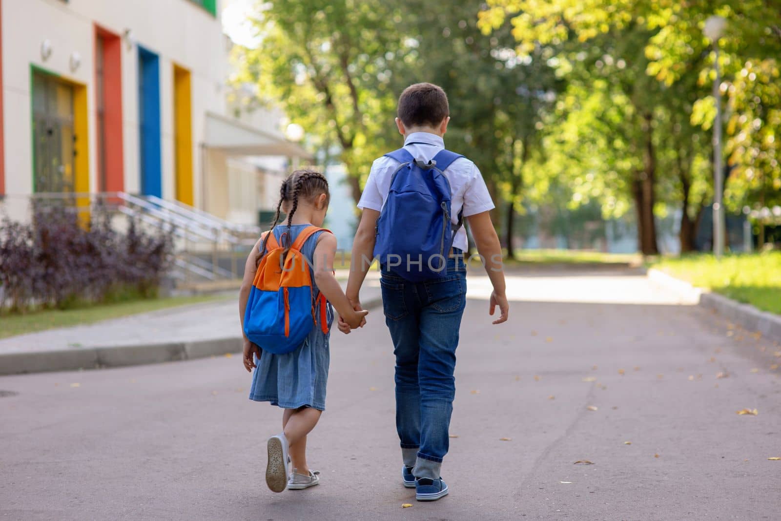 schoolchildren, a little girl and a boy, brother and sister with backpacks hold hands go to school with multi-colored windows and green trees in the morning on a sunny summer day. Close up. Mock up