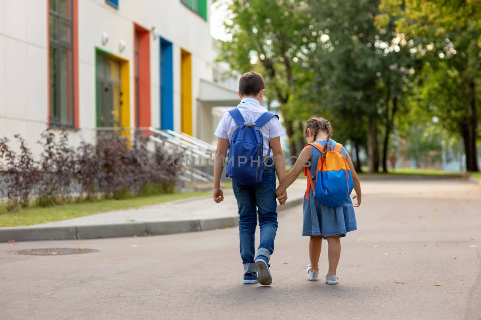 two schoolchildren, brother and sister with backpacks hold hands go to school in the morning on a sunny summer day. Mock up. Copy space