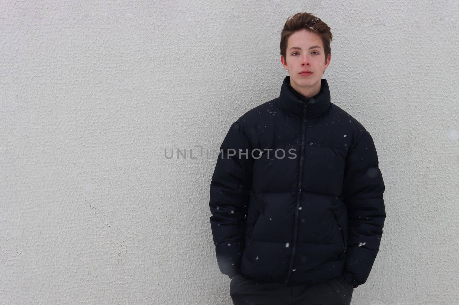 Seriously handsome teenage boy looking at the camera and holding his hands in his pockets against a backdrop of falling snowflakes. by gelog67