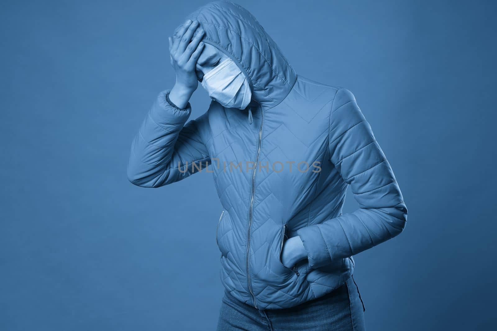 Young brunette woman in protective face mask and coat over blue background. Model girl in medical mask and spring outfit, studio shot with copy space for your text. Anti-flu, anti-epidemic