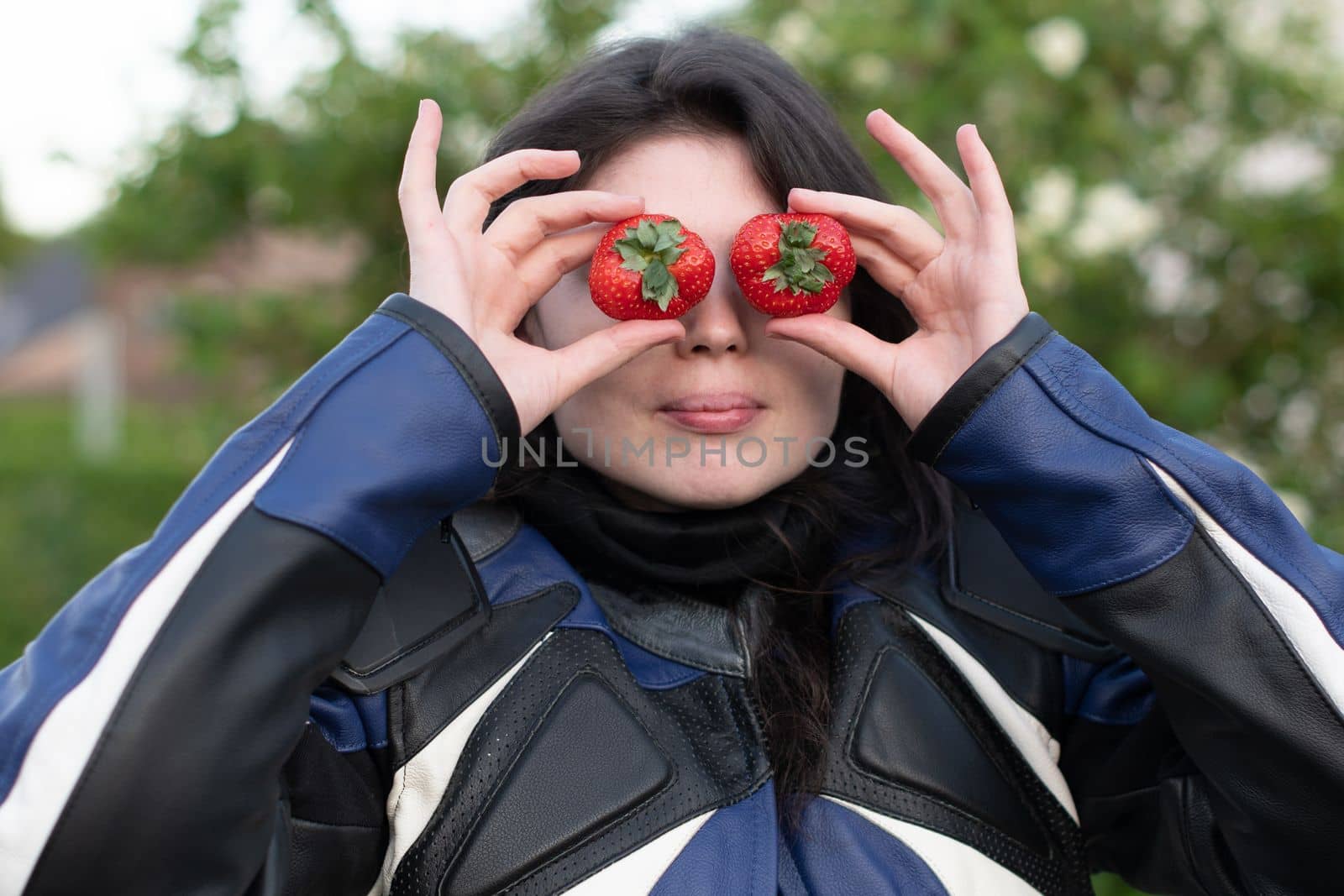 beautiful young woman motobiker in black leather pants, black boots and a black helmet, in a blue jacket on a red honda motorcycle, put strawberries to her eyes. High quality photo