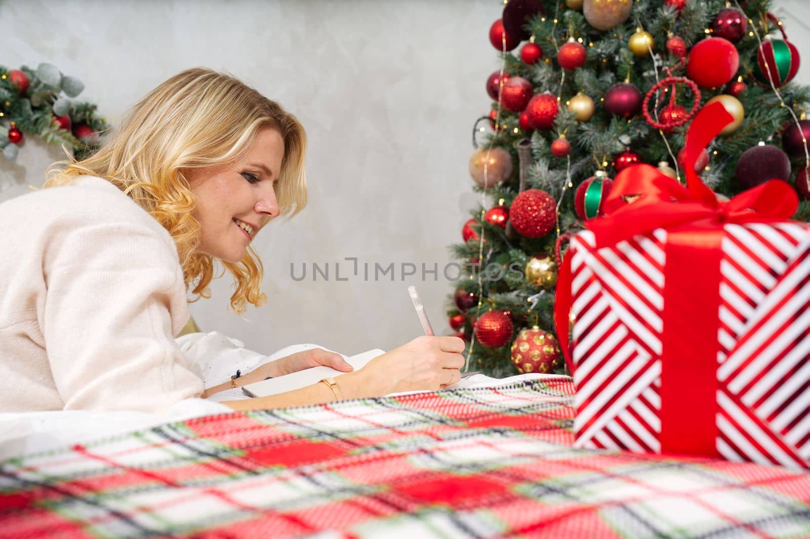 happy woman with pen and notebook making wish list or to do list for new year in bed over christmas tree. woman dreaming in christmas time. xmas holidays concept