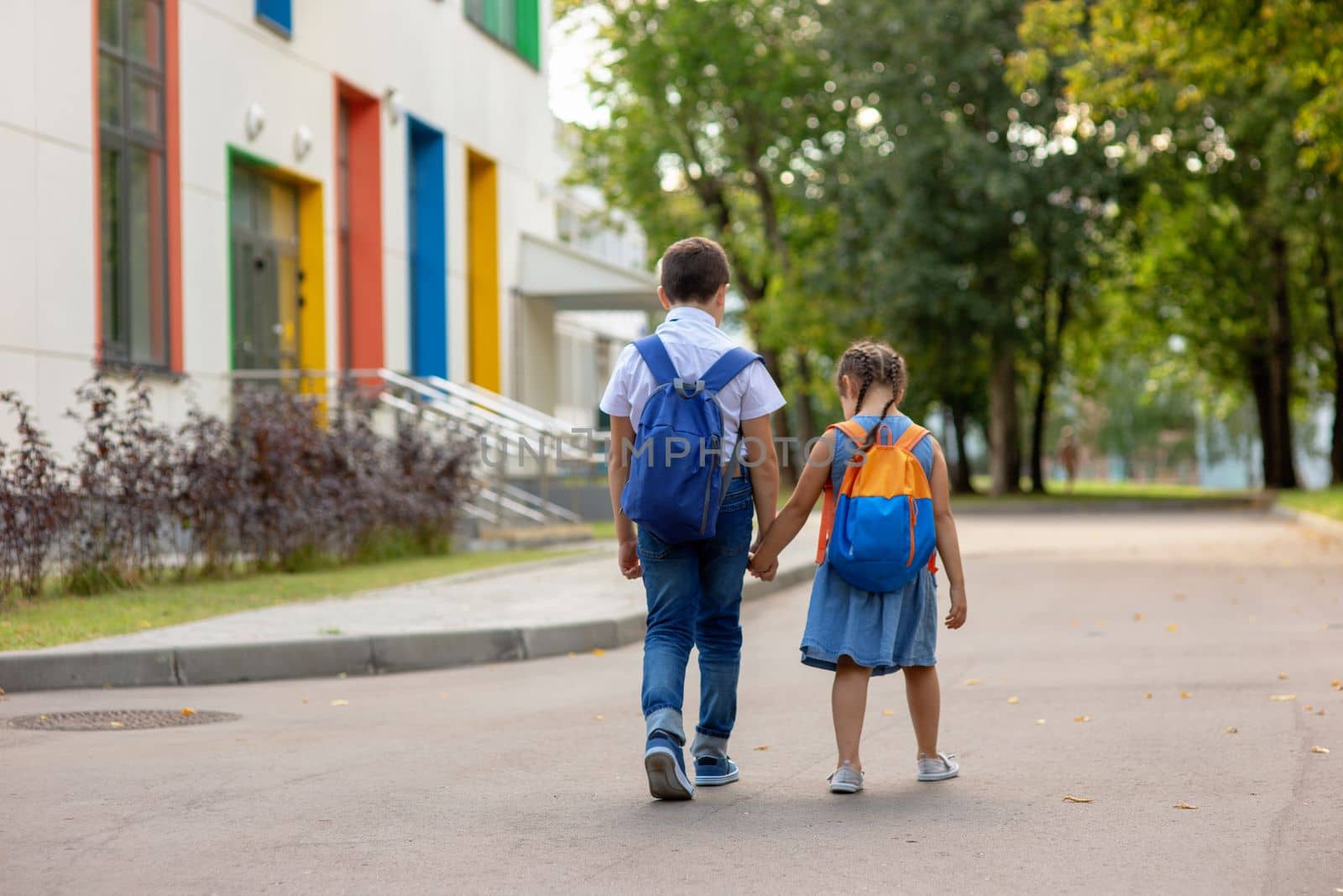 two schoolchildren, brother and sister with backpacks hold hands go to school in the morning on a sunny summer day on background school bulding . Mock up. Copy space