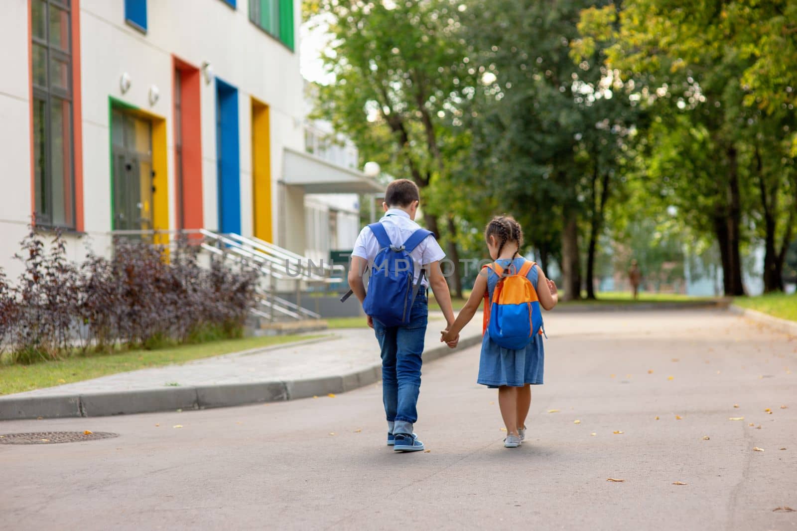 two schoolchildren, a little girl and a boy, brother and sister with backpacks hold hands go to school in the morning on a sunny summer day. Mock up. Copy space