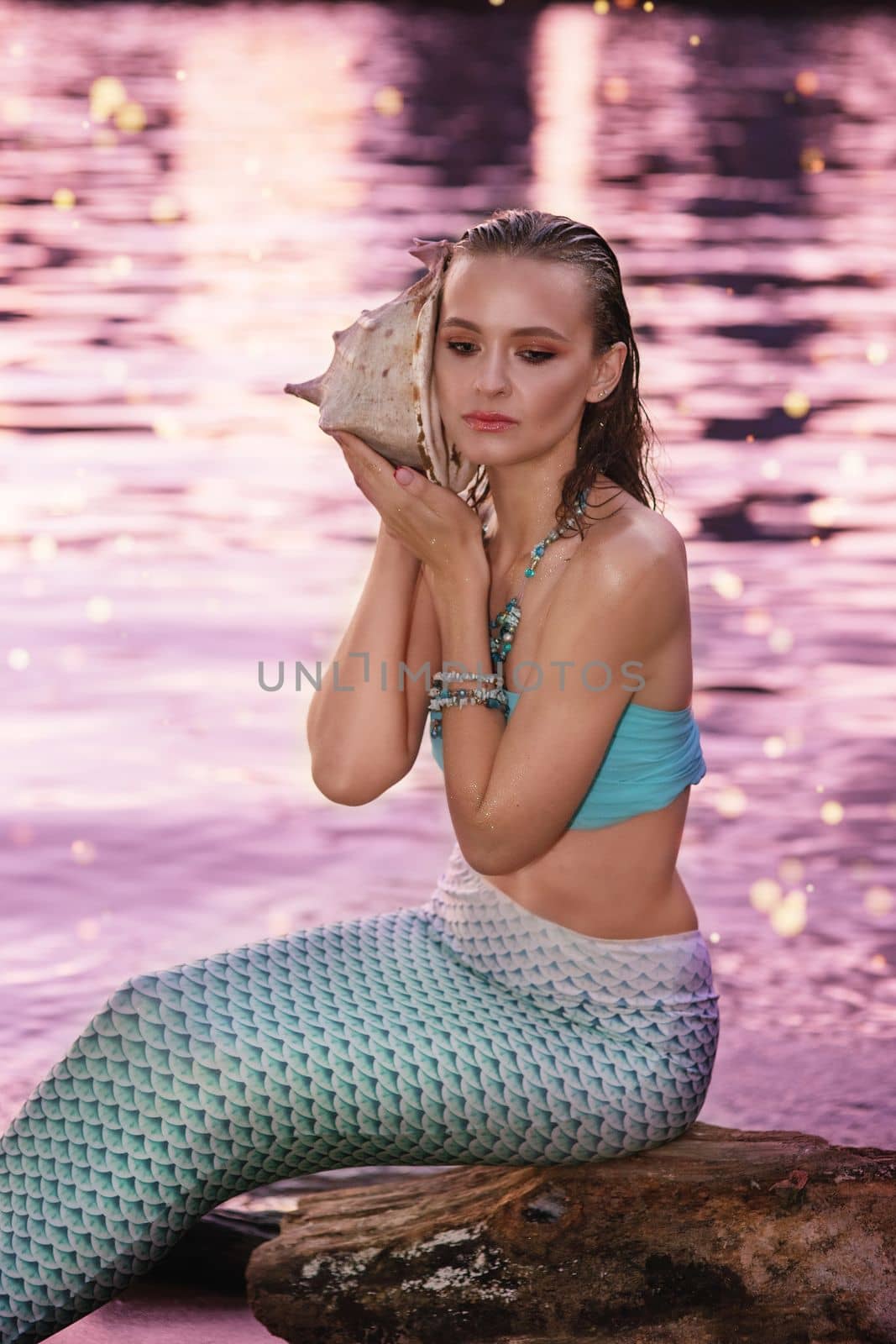 A mermaid girl sits near the water at dusk, holding a large seashell.