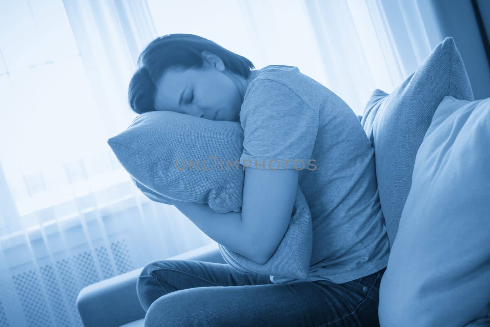 Sad depressed woman at home, she is sitting on the couch and hugging a pillow, loneliness and sadness concept
