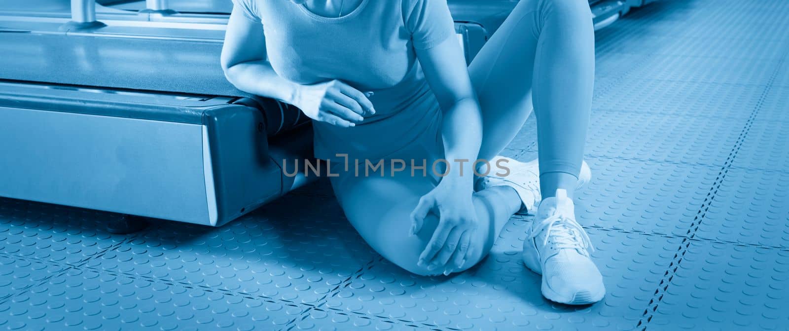 Young woman in sportswear having pain in her knee while training in gym, Girl sitting on a floor touching her knee in pain by Mariakray