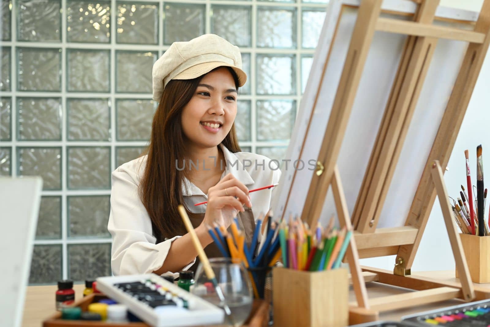Smiling female painter sitting in front of canvas and painting picture with watercolor. Leisure activity concept.