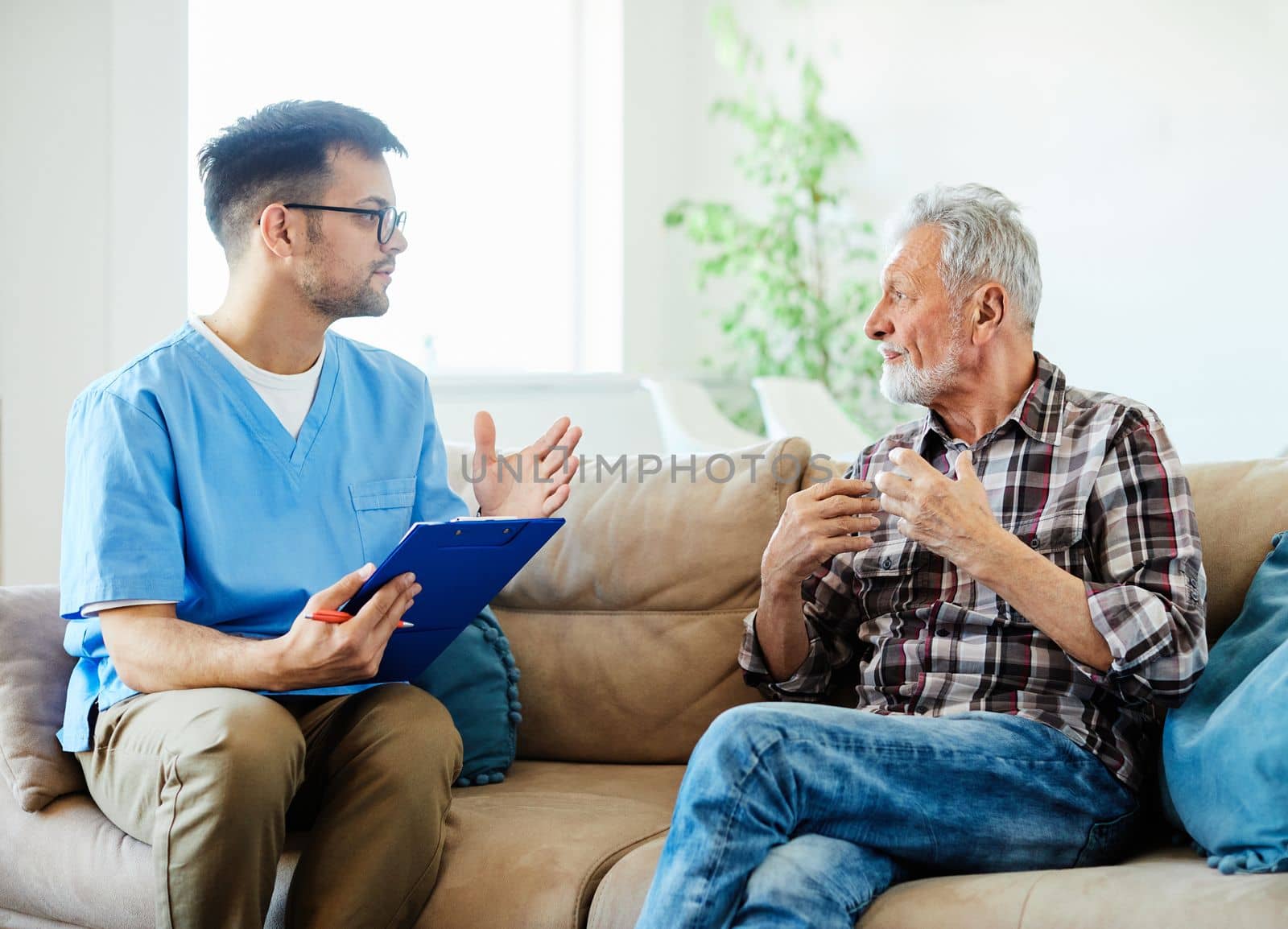 Doctor or nurse caregiver talking with senior man on sofa at home or nursing home