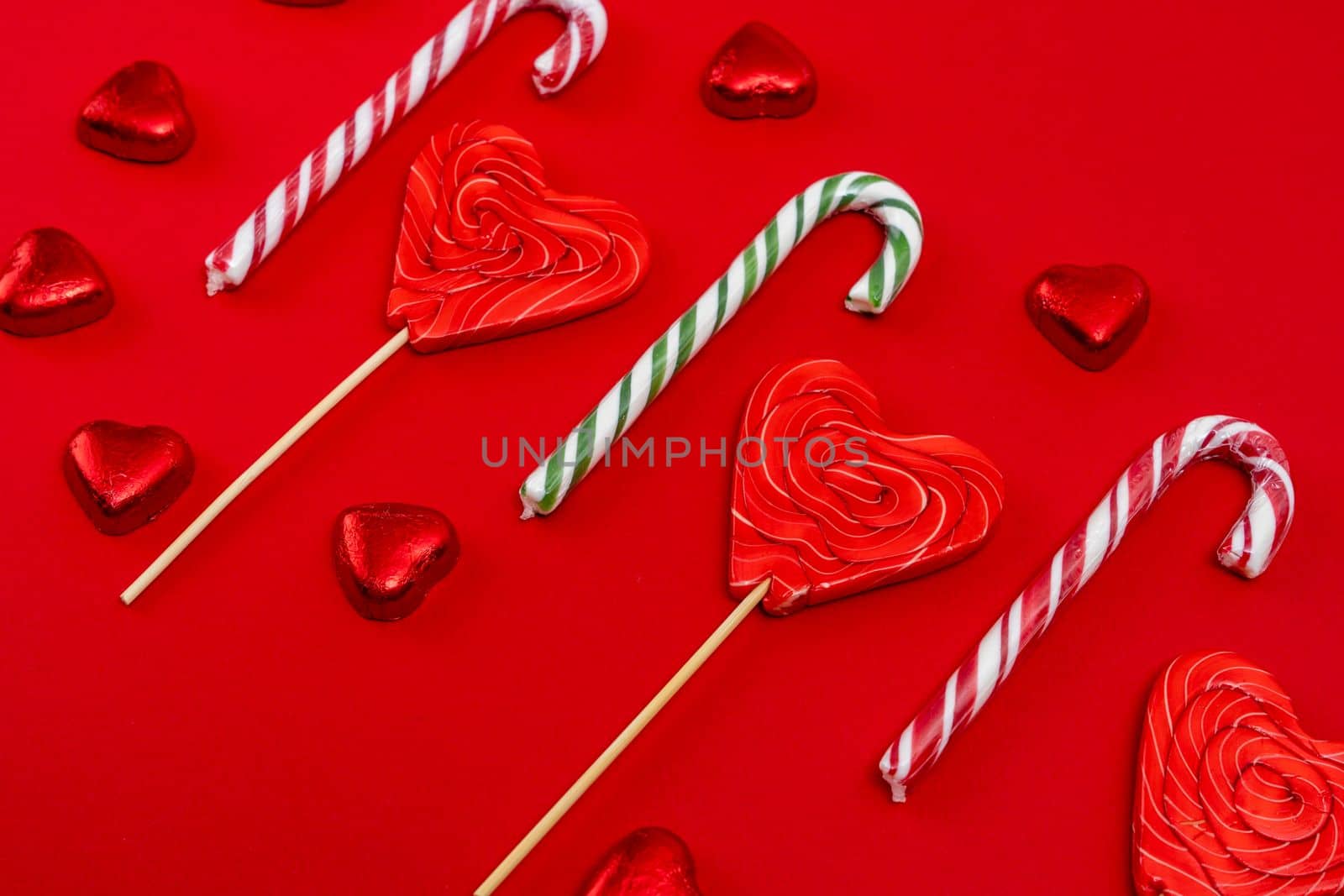 red festive Christmas background with candy in the shape of hearts wrapped in shiny foil and candy in the shape of a caramel cane